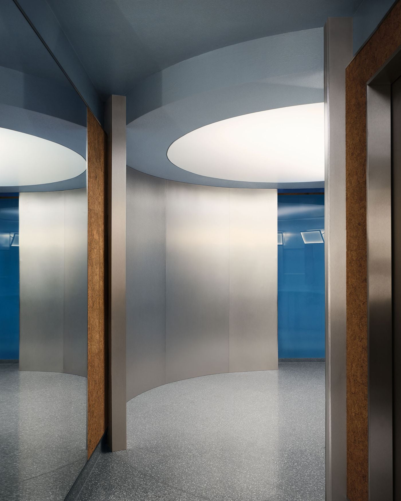 A circulation area combines curved metallic partitions, cork-clad wall panels and a speckled grey floor beneath oversized circular ceiling lights. Cool blue walls and glimpses of brighter blue beyond reinforce the women’s changing-area palette, while the rounded geometry gives the corridor a distinctly retro-futurist quality. A circulation area combines curved metallic partitions, cork-clad wall panels and a speckled grey floor beneath oversized circular ceiling lights. Cool blue walls and glimpses of brighter blue beyond reinforce the women’s changing-area palette, while the rounded geometry gives the corridor a distinctly retro-futurist quality.