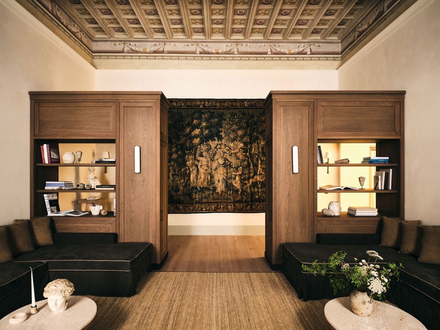 Beneath a painted coffered ceiling with rosettes and garlanded friezes, two oak bookcase towers frame an antique tapestry of classical figures in a wooded landscape at La Réserve Firenze. Deep brown banquettes with contrast piping, round stone-topped tables and shelves of books, ceramics and fragments give the lounge a scholarly calm. Beneath a painted coffered ceiling with rosettes and garlanded friezes, two oak bookcase towers frame an antique tapestry of classical figures in a wooded landscape at La Réserve Firenze. Deep brown banquettes with contrast piping, round stone-topped tables and shelves of books, ceramics and fragments give the lounge a scholarly calm.