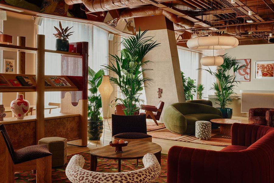 Holloway Li breaks the expansive ground floor into intimate lounge settings using bespoke joinery, plants, rugs and sculptural seating. Burgundy, olive and patterned upholstery animate the warm timber interior, while angular concrete columns and exposed ceiling services provide a raw architectural counterpoint to the mid-century-inspired furnishings.