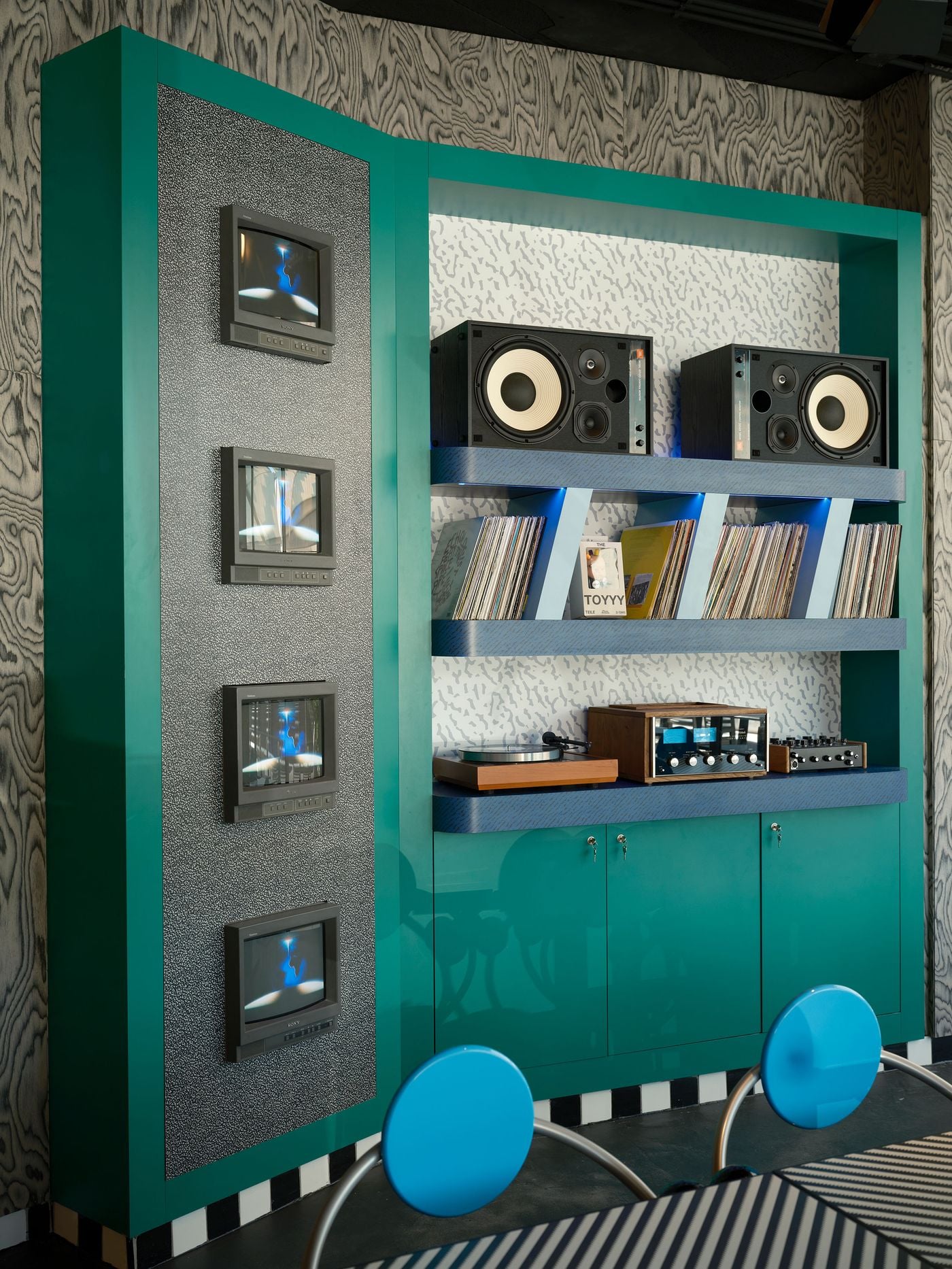 A close-up of the teal-lacquered media wall at Seth & Manni's shows four vintage Sony monitors set into a speckled grey panel. Blue patterned shelves hold JBL speakers, vinyl records, a turntable and a vintage amplifier against squiggle-patterned backing. The ensemble recalls the founders' lives behind the decks. A close-up of the teal-lacquered media wall at Seth & Manni's shows four vintage Sony monitors set into a speckled grey panel. Blue patterned shelves hold JBL speakers, vinyl records, a turntable and a vintage amplifier against squiggle-patterned backing. The ensemble recalls the founders' lives behind the decks.