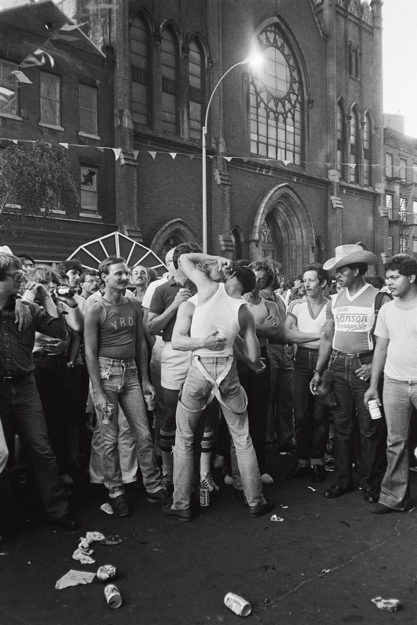 Before the brick facade of a Gothic Revival church, its rose window glowing under a lit streetlamp, two men lock in a kiss at the centre of a crowd. Onlookers in tank tops, denim and a straw cowboy hat watch, beer cans in hand, as litter scatters across the street in this charged image from Don't Worry Boys. Before the brick facade of a Gothic Revival church, its rose window glowing under a lit streetlamp, two men lock in a kiss at the centre of a crowd. Onlookers in tank tops, denim and a straw cowboy hat watch, beer cans in hand, as litter scatters across the street in this charged image from Don't Worry Boys.