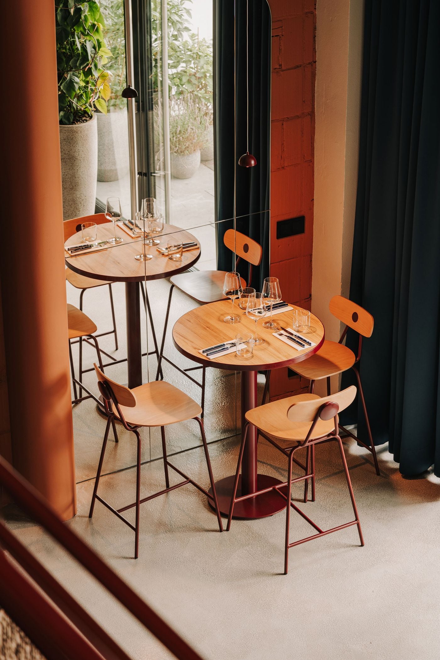 An intimate dining nook at WIN wine bar in Warsaw features a round timber-topped table paired with bentwood stools on slender burgundy metal frames. A floor-to-ceiling mirror embedded in the wall reflects the table and its surroundings, creating the illusion of a larger space, while terracotta masonry, deep blue curtains and miniature pendant lights complete the composition. An intimate dining nook at WIN wine bar in Warsaw features a round timber-topped table paired with bentwood stools on slender burgundy metal frames. A floor-to-ceiling mirror embedded in the wall reflects the table and its surroundings, creating the illusion of a larger space, while terracotta masonry, deep blue curtains and miniature pendant lights complete the composition.