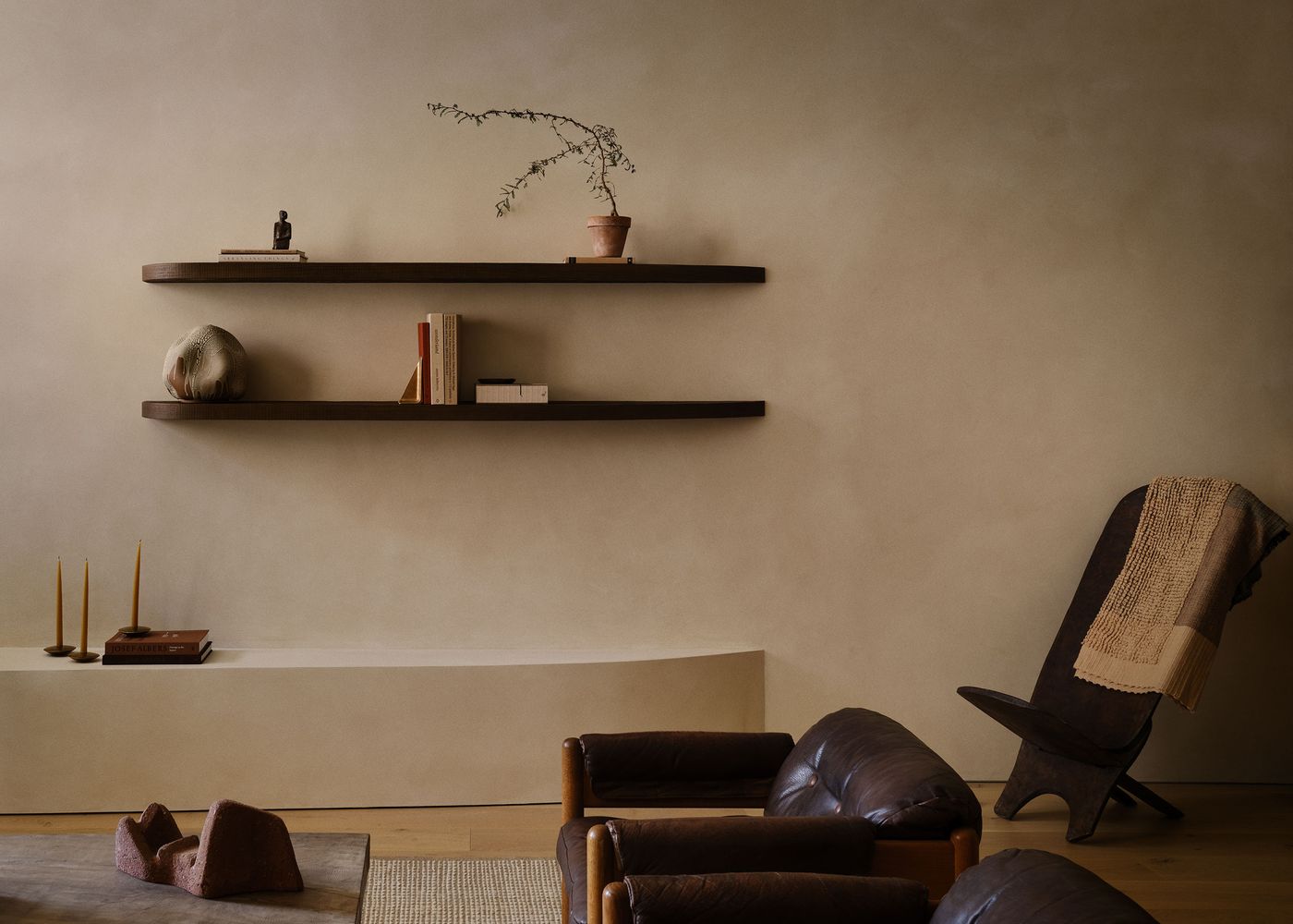 A minimalistic living space featuring brown leather chairs, wooden shelves with books and a plant, and a textured beige wall. A minimalistic living space featuring brown leather chairs, wooden shelves with books and a plant, and a textured beige wall.