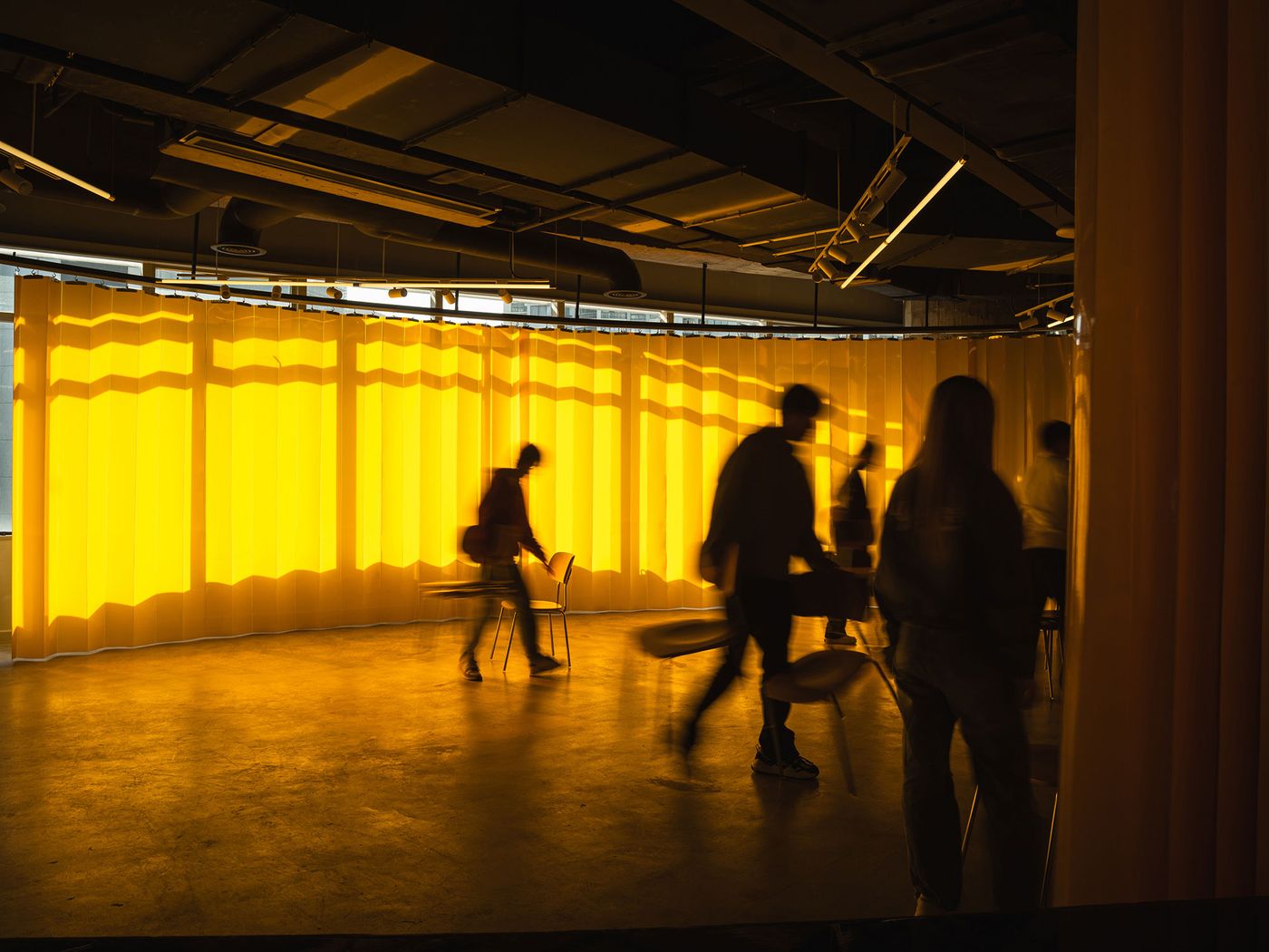Silhouetted figures move against a luminous, floor-to-ceiling yellow curtain in 889 GLO Art Space, creating a dramatic, artful scene. The golden light spilling onto the polished concrete floor enhances the atmospheric mood, transforming a functional partition into a dynamic, artistic backdrop within the industrial setting. Silhouetted figures move against a luminous, floor-to-ceiling yellow curtain in 889 GLO Art Space, creating a dramatic, artful scene. The golden light spilling onto the polished concrete floor enhances the atmospheric mood, transforming a functional partition into a dynamic, artistic backdrop within the industrial setting.