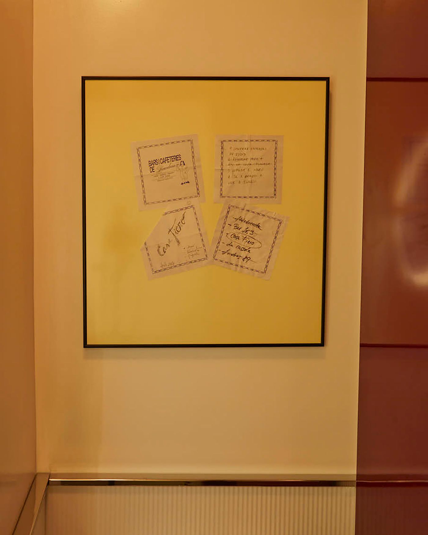 A square, framed piece of art hangs against a warm-toned wall. The artwork itself is a simple, minimalist composition of four handwritten notes on pale paper, arranged askew on a solid yellow background. The black frame and simple content emphasize a subtle, personal sense of artistry.