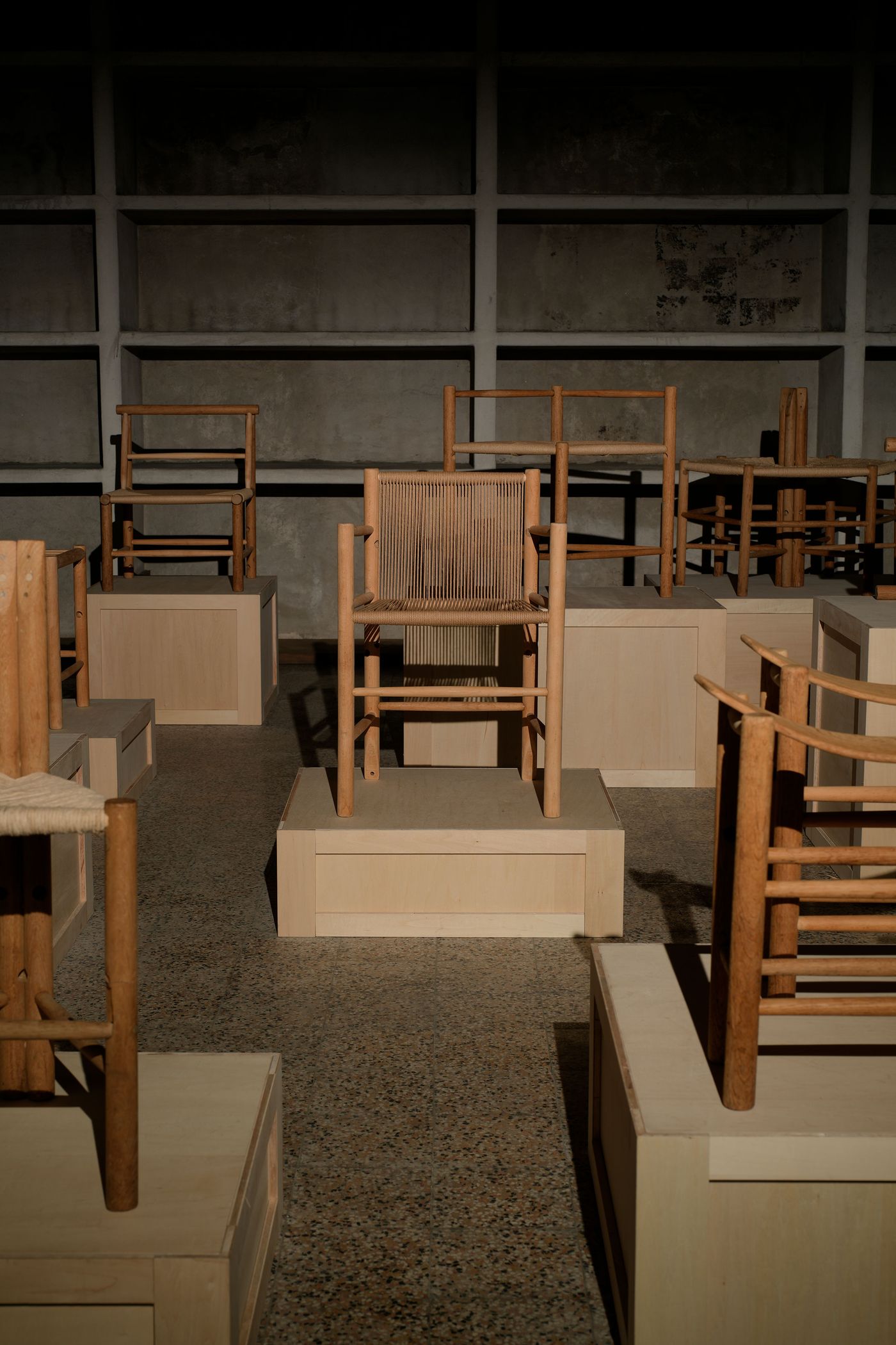A dimly lit showcase features various wooden chairs displayed on light-colored pedestals against a textured dark wall.