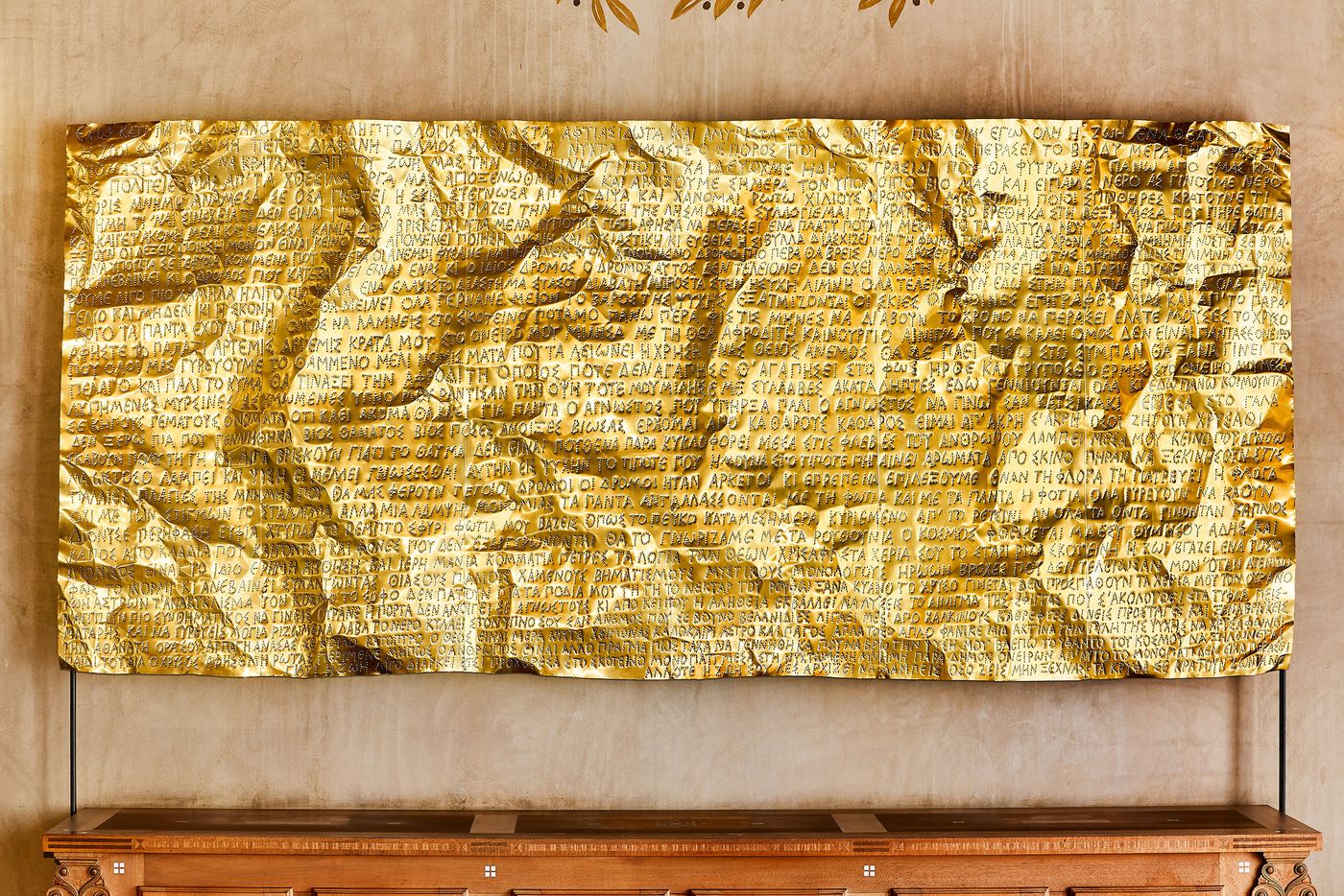 A focused shot of Gabriel Leger's installation, a large rectangular sheet of gold-colored metal. The material is deliberately crinkled and textured, with Greek text subtly etched across its surface, giving it the appearance of a weighty, ancient manuscript reinterpreted for a contemporary space. A focused shot of Gabriel Leger's installation, a large rectangular sheet of gold-colored metal. The material is deliberately crinkled and textured, with Greek text subtly etched across its surface, giving it the appearance of a weighty, ancient manuscript reinterpreted for a contemporary space.