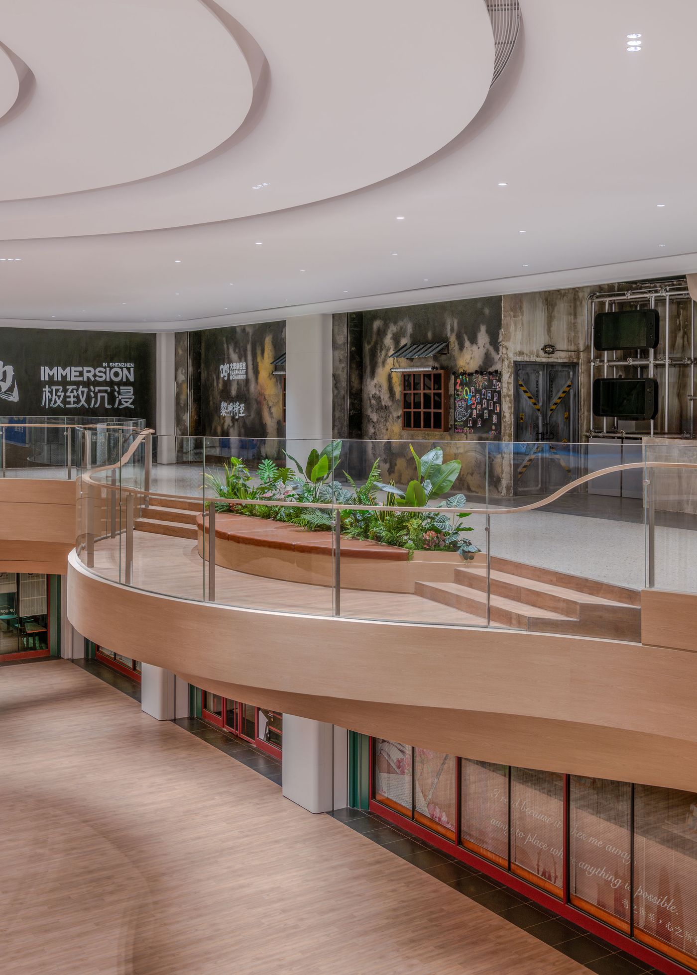 A modern interior view showcasing a curved wooden railing, greenery, and artistic wall murals in a spacious, well-lit atrium.