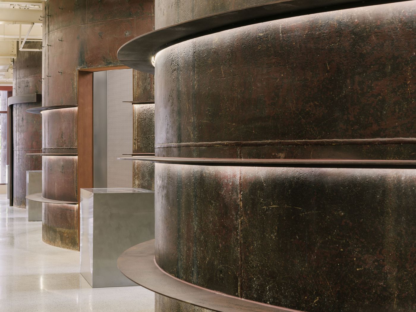 A dramatic perspective highlights the majestic, curved walls of patinated steel, featuring subtle integrated lighting along their horizontal bands, emphasizing their monumental presence. These industrial forms are juxtaposed with a sleek, minimalist grey plinth and a finely perforated metal screen, creating a compelling dialogue between raw and refined textures. A dramatic perspective highlights the majestic, curved walls of patinated steel, featuring subtle integrated lighting along their horizontal bands, emphasizing their monumental presence. These industrial forms are juxtaposed with a sleek, minimalist grey plinth and a finely perforated metal screen, creating a compelling dialogue between raw and refined textures.
