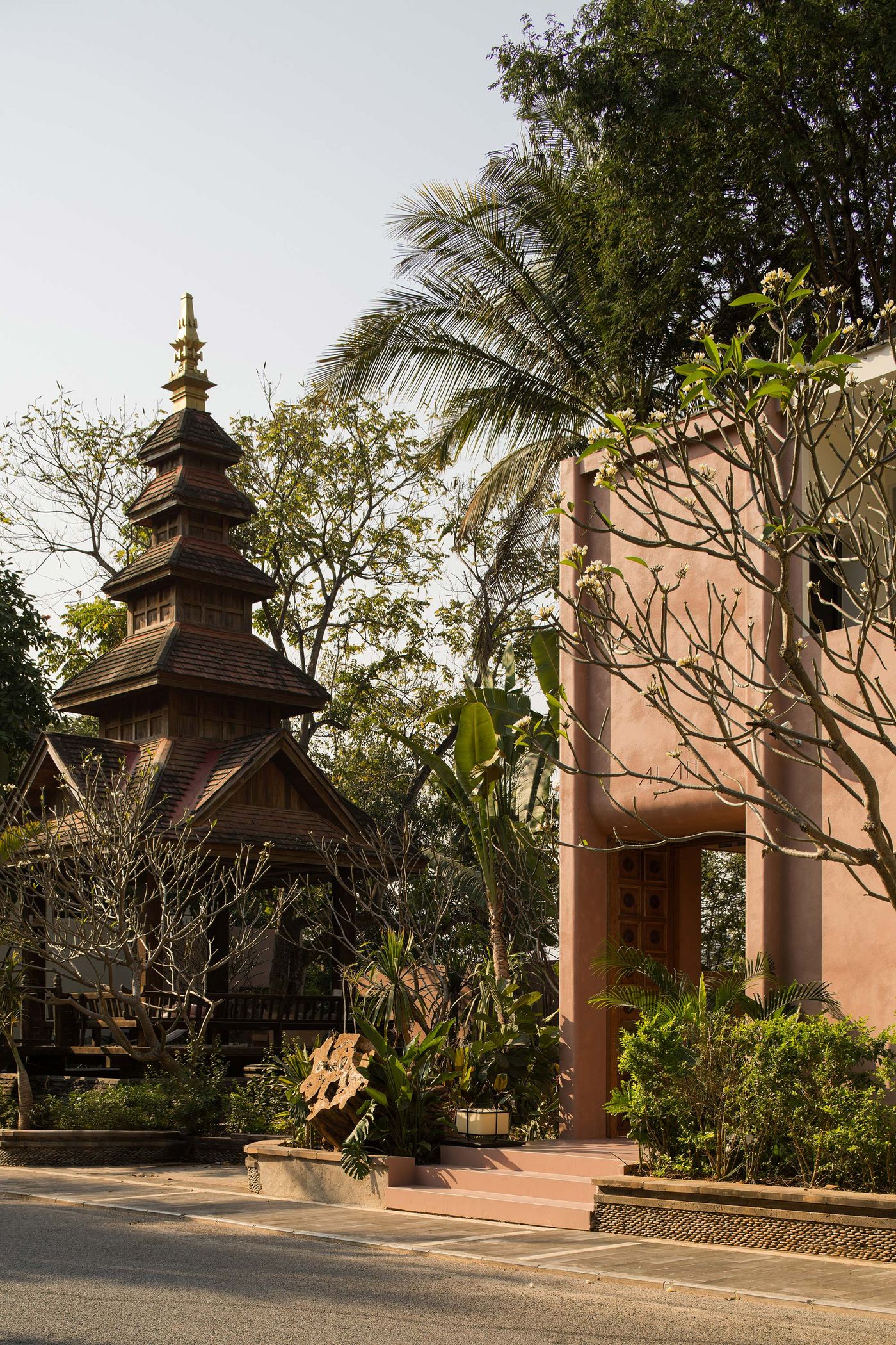 A wooden temple with a tiered roof stands beside a pink structure, surrounded by tropical plants and trees under a clear sky. A wooden temple with a tiered roof stands beside a pink structure, surrounded by tropical plants and trees under a clear sky.