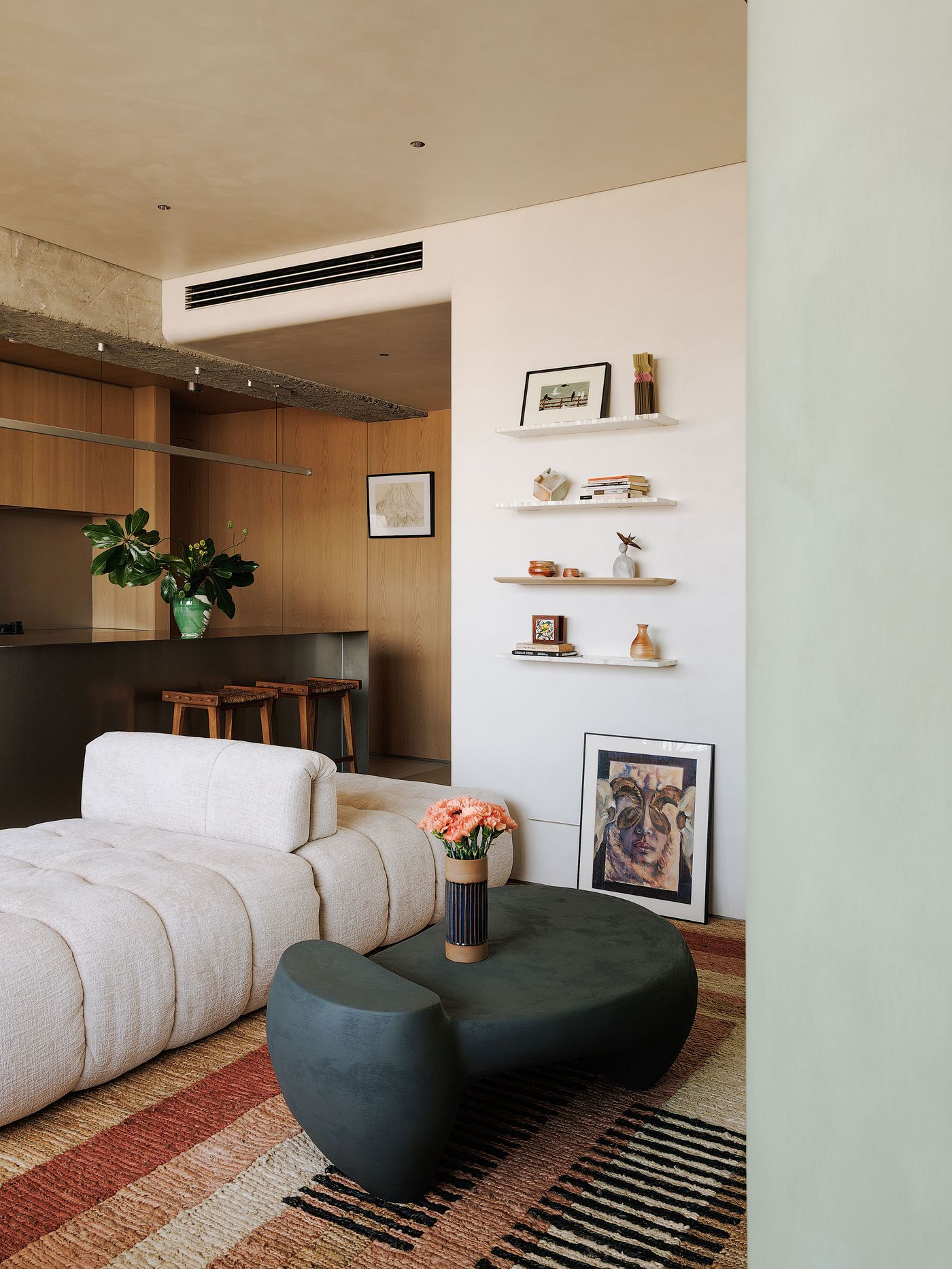 A contemporary living space imbued with eclectic details. A plush cream modular sofa sits on a geometric patterned rug in earthy tones, facing a sculptural dark gray coffee table adorned with flowers. Floating white shelves display curated objects and art, while the kitchen’s wooden cabinetry and a concrete beam provide a backdrop, showcasing a blend of textures and artful arrangements. A contemporary living space imbued with eclectic details. A plush cream modular sofa sits on a geometric patterned rug in earthy tones, facing a sculptural dark gray coffee table adorned with flowers. Floating white shelves display curated objects and art, while the kitchen’s wooden cabinetry and a concrete beam provide a backdrop, showcasing a blend of textures and artful arrangements.