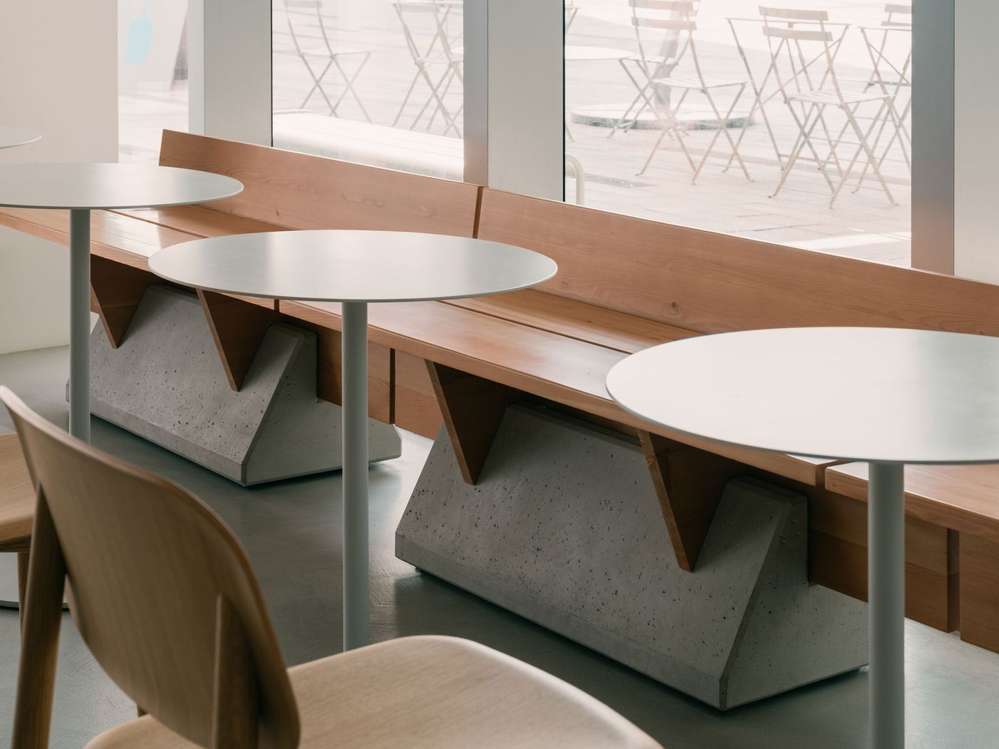Angular timber seating mounted on bold concrete bases forms a custom bench by the window, blending craftsmanship with brutalist pragmatism. Pale grey tables and chairs continue the interior’s sculptural restraint.