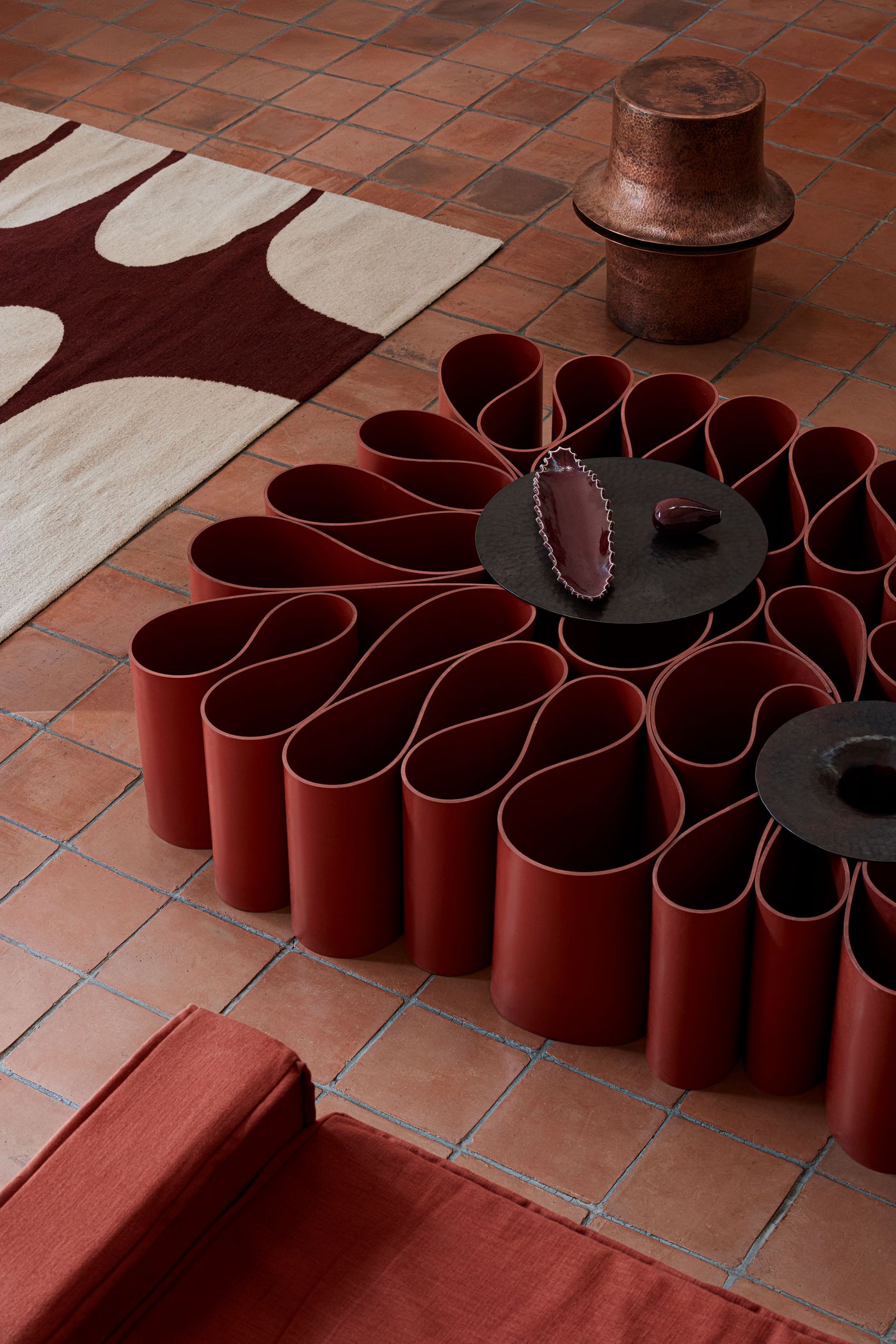 A modern living space featuring a red, flower-shaped coffee table, terra cotta tiles, a patterned rug, and stylish decor accents.
