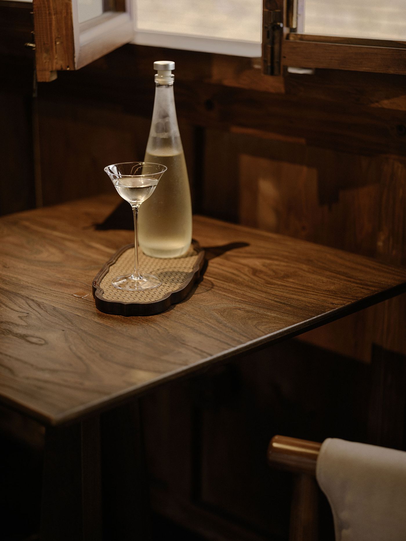 A crystal cocktail glass with clear drink sits on a woven tray beside a bottle, all arranged on a polished wooden table. A crystal cocktail glass with clear drink sits on a woven tray beside a bottle, all arranged on a polished wooden table.