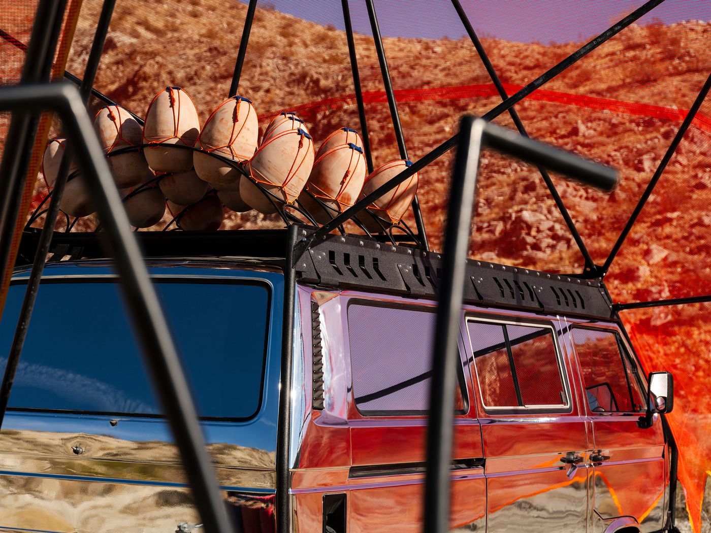 A shiny van with orange buoy-like structures on its roof, surrounded by a geometric frame and a rocky landscape in the background.
