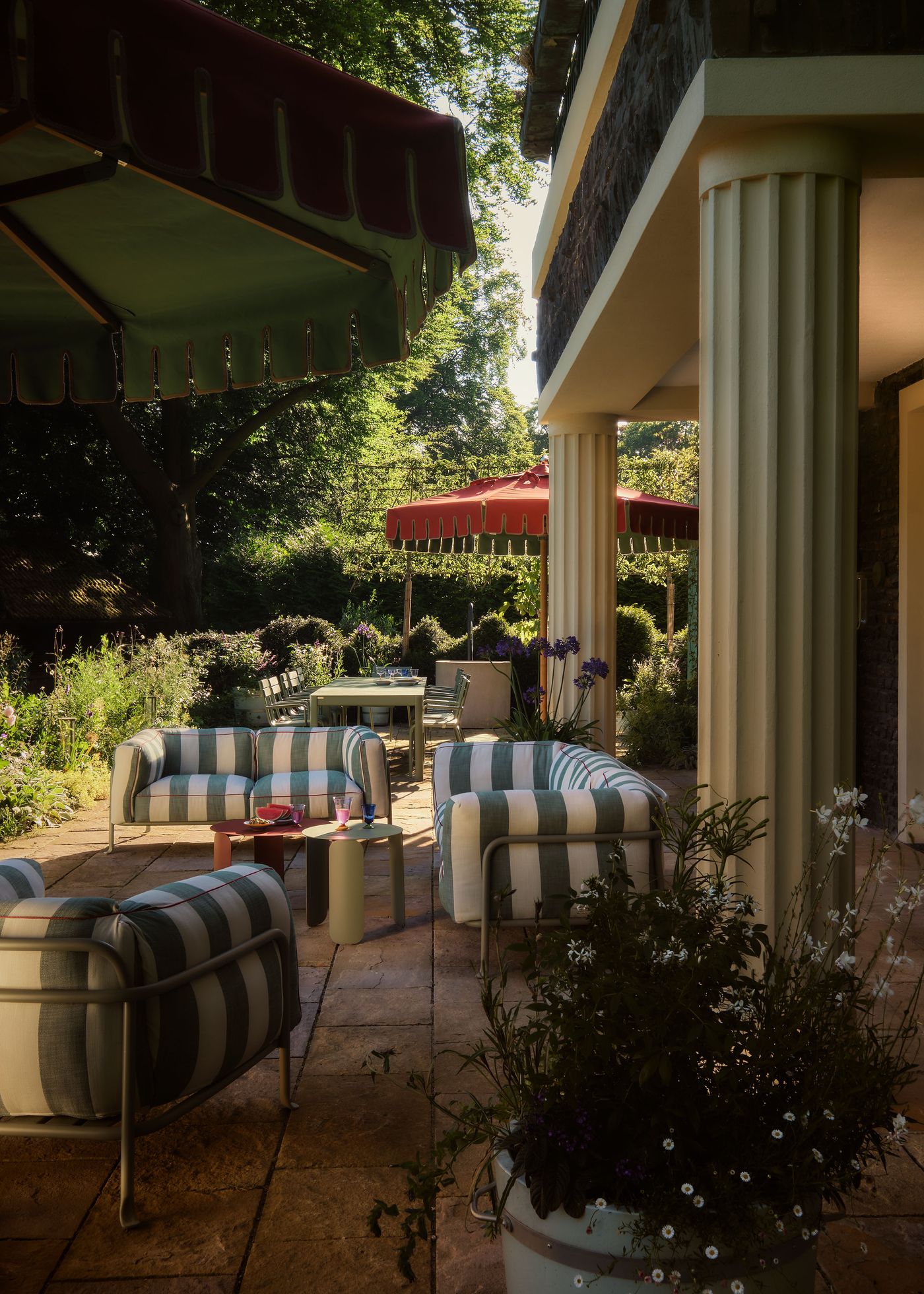 A cozy outdoor patio with striped chairs, colorful tables, lush greenery, and shaded by large awnings, inviting relaxation and gatherings.