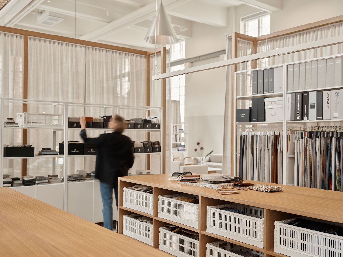 A creative workspace features a long wooden table with organized white baskets for materials, and tall white shelving units displaying various design samples and fabric swatches. A person is seen organizing items, highlighting the functional and organized nature of the studio. A creative workspace features a long wooden table with organized white baskets for materials, and tall white shelving units displaying various design samples and fabric swatches. A person is seen organizing items, highlighting the functional and organized nature of the studio.