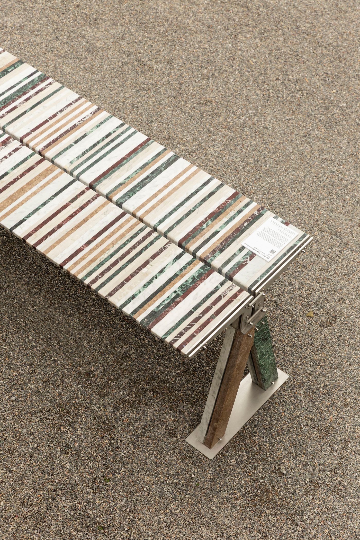 An abstract outdoor bench is crafted from a mosaic of rectangular marble tiles, each showcasing distinct stripes of burgundy, green, and beige. The top is supported by angular legs, one of which is a combination of stone and wood, set on a metal base. The composition emphasizes the intricate patterns and rich textures of the natural materials against the gravelly ground, highlighting a modern, sculptural approach to furniture design.