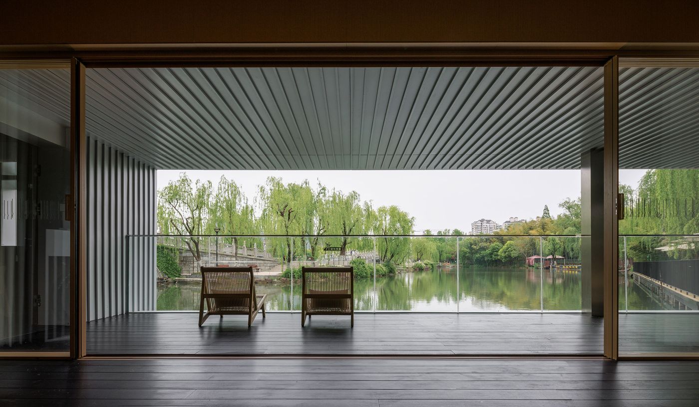 From within Wutopia Lab's Lake House, a serene vista unfolds through expansive sliding glass doors that open onto a narrow balcony. Two minimalist wooden chairs face the tranquil lake, framed by the lush green of weeping willows. The dark wooden floor and simple aesthetic create a calm, Japandi-inspired interior, drawing the eye towards the immersive natural panorama beyond. From within Wutopia Lab's Lake House, a serene vista unfolds through expansive sliding glass doors that open onto a narrow balcony. Two minimalist wooden chairs face the tranquil lake, framed by the lush green of weeping willows. The dark wooden floor and simple aesthetic create a calm, Japandi-inspired interior, drawing the eye towards the immersive natural panorama beyond.