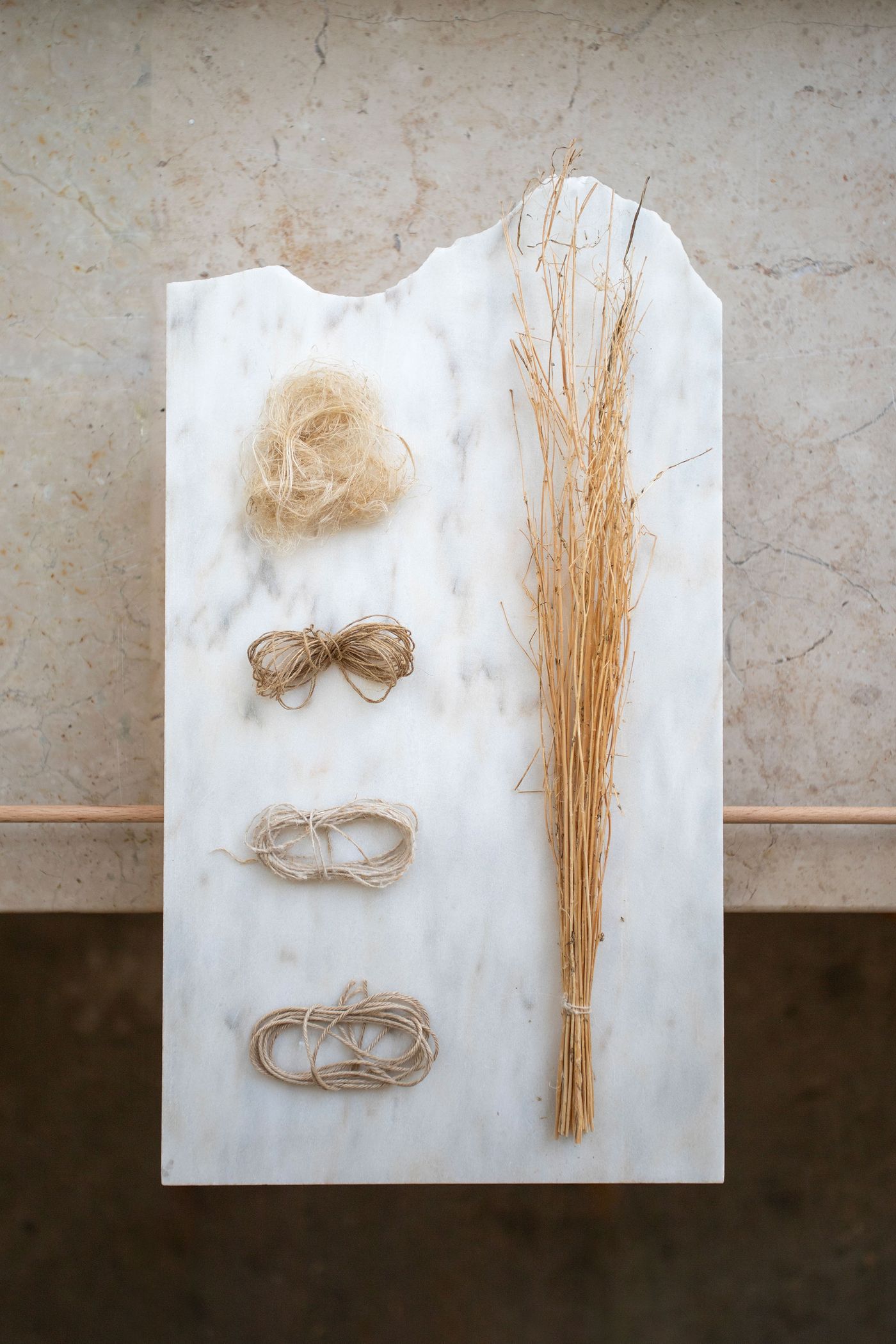 A pristine white marble board becomes a canvas for natural materials, displaying various hemp fibers and dried grasses in an orderly composition. The weathered stone backdrop and warm wooden ledge frame this study in texture, reflecting a biophilic approach to material exploration and craft fundamentals.
