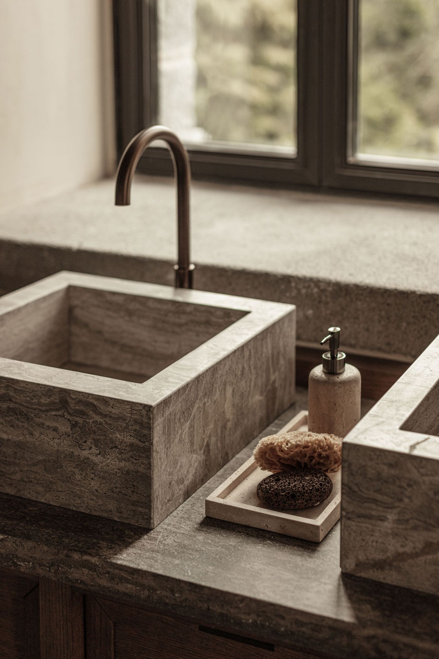 A close-up reveals exquisite material selection, featuring twin rectangular vessel sinks crafted from veined grey stone. A brushed bronze faucet adds a touch of understated luxury, while natural sponges on a stone tray emphasize a biophilic and mindful approach to daily rituals, set against a backdrop of a framed window and stone ledge. A close-up reveals exquisite material selection, featuring twin rectangular vessel sinks crafted from veined grey stone. A brushed bronze faucet adds a touch of understated luxury, while natural sponges on a stone tray emphasize a biophilic and mindful approach to daily rituals, set against a backdrop of a framed window and stone ledge.
