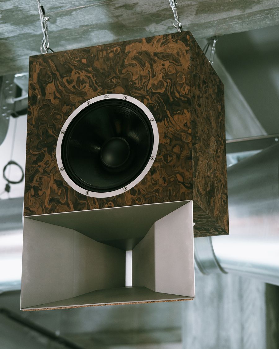 A stylish wooden speaker with a black cone and metallic horn, suspended from a ceiling in an industrial space. A stylish wooden speaker with a black cone and metallic horn, suspended from a ceiling in an industrial space.