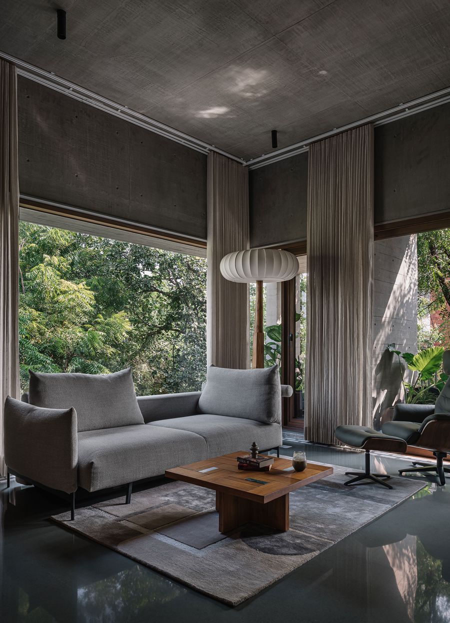 A spacious living area featuring a minimalist grey sofa and a classic Eames Lounge Chair, set against a backdrop of floor-to-ceiling windows that offer a lush, biophilic view of dense trees. The raw concrete ceiling and dark polished floor are softened by a large abstract rug and a striking pleated pendant light, blending brutalist elements with refined comfort.
