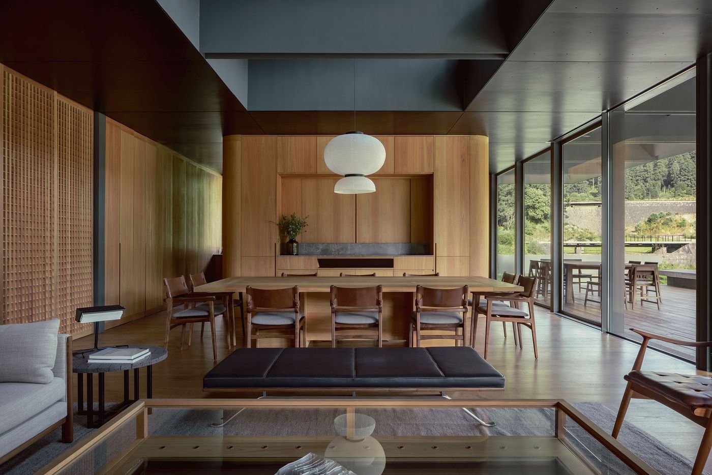 A modern, warm wood dining room featuring a long table, stylish chairs, and large windows with a natural view. Minimalist decor and soft lighting.