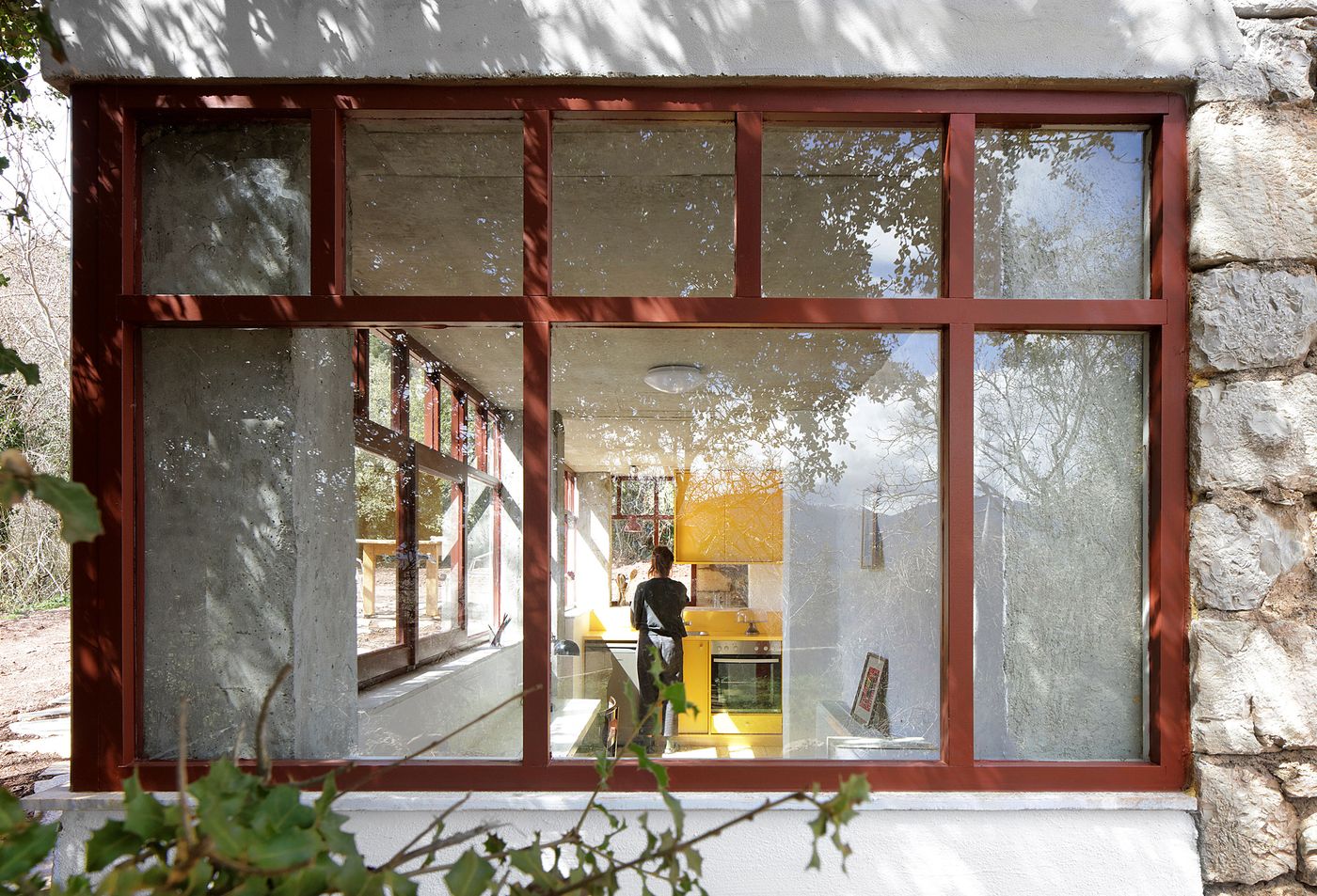 A figure moves through the transparent kitchen pavilion, where coral-red steel framing creates architectural rhythm against whitewashed stone walls. The contemporary intervention transforms daily rituals into a theatrical performance, visible through floor-to-ceiling glazing that dissolves interior boundaries.
