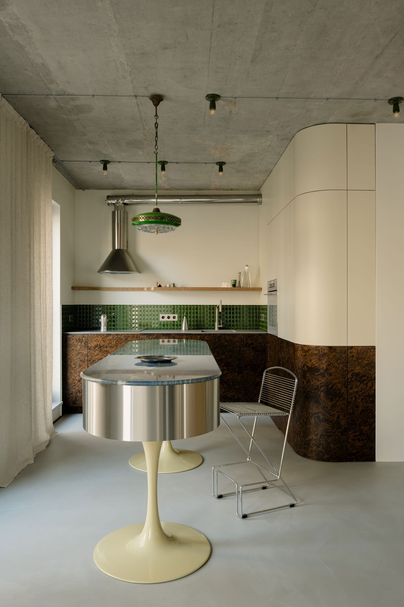 This wide-angle kitchen view reveals the fluid geometry of the custom island and cabinetry, where 1970s influences meet minimalism through green glazed tiles, curved cream panels, and richly figured wood veneer. This wide-angle kitchen view reveals the fluid geometry of the custom island and cabinetry, where 1970s influences meet minimalism through green glazed tiles, curved cream panels, and richly figured wood veneer.