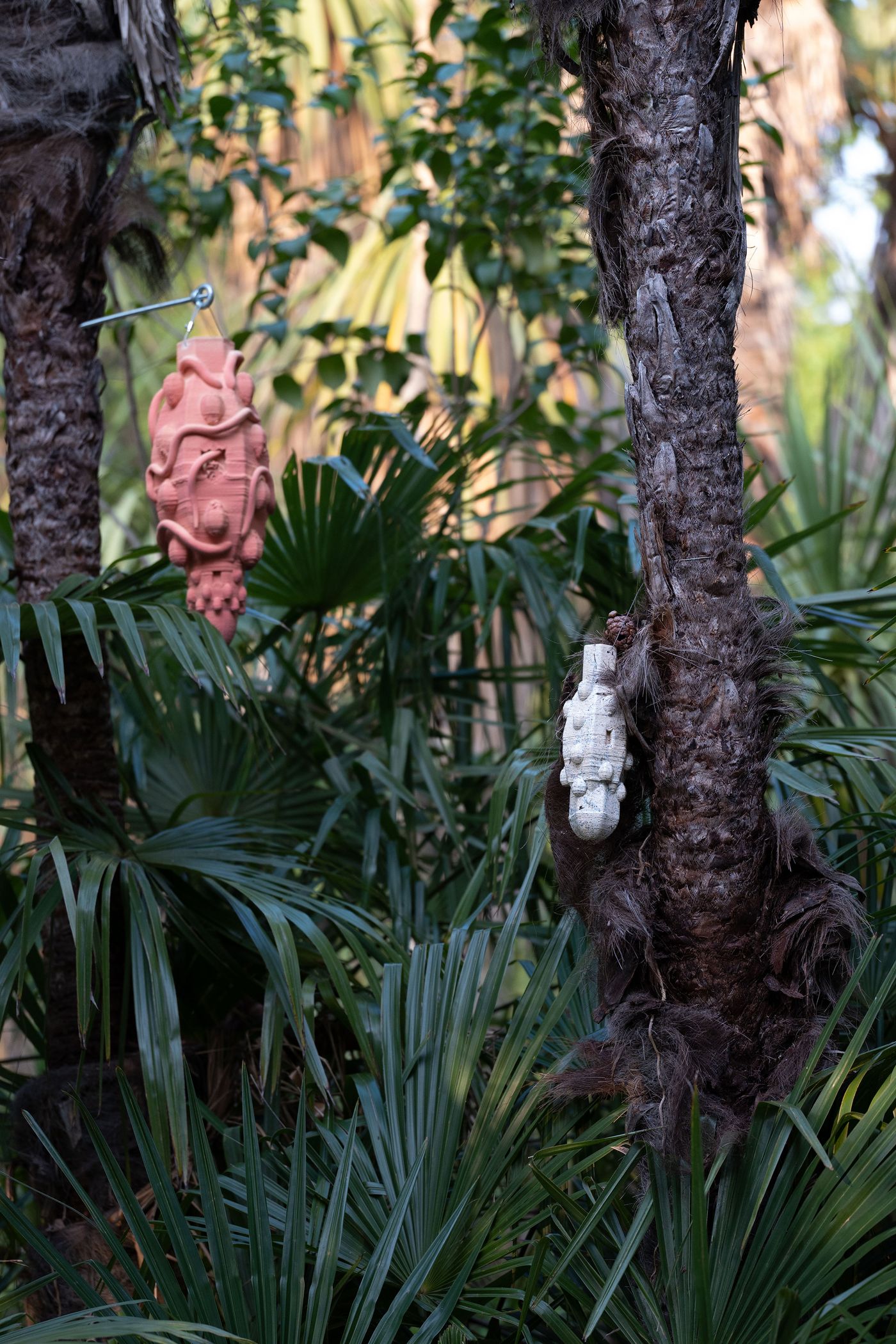 Two abstract sculptures, one pink and one white, hang from palm trees amidst lush green foliage. Two abstract sculptures, one pink and one white, hang from palm trees amidst lush green foliage.