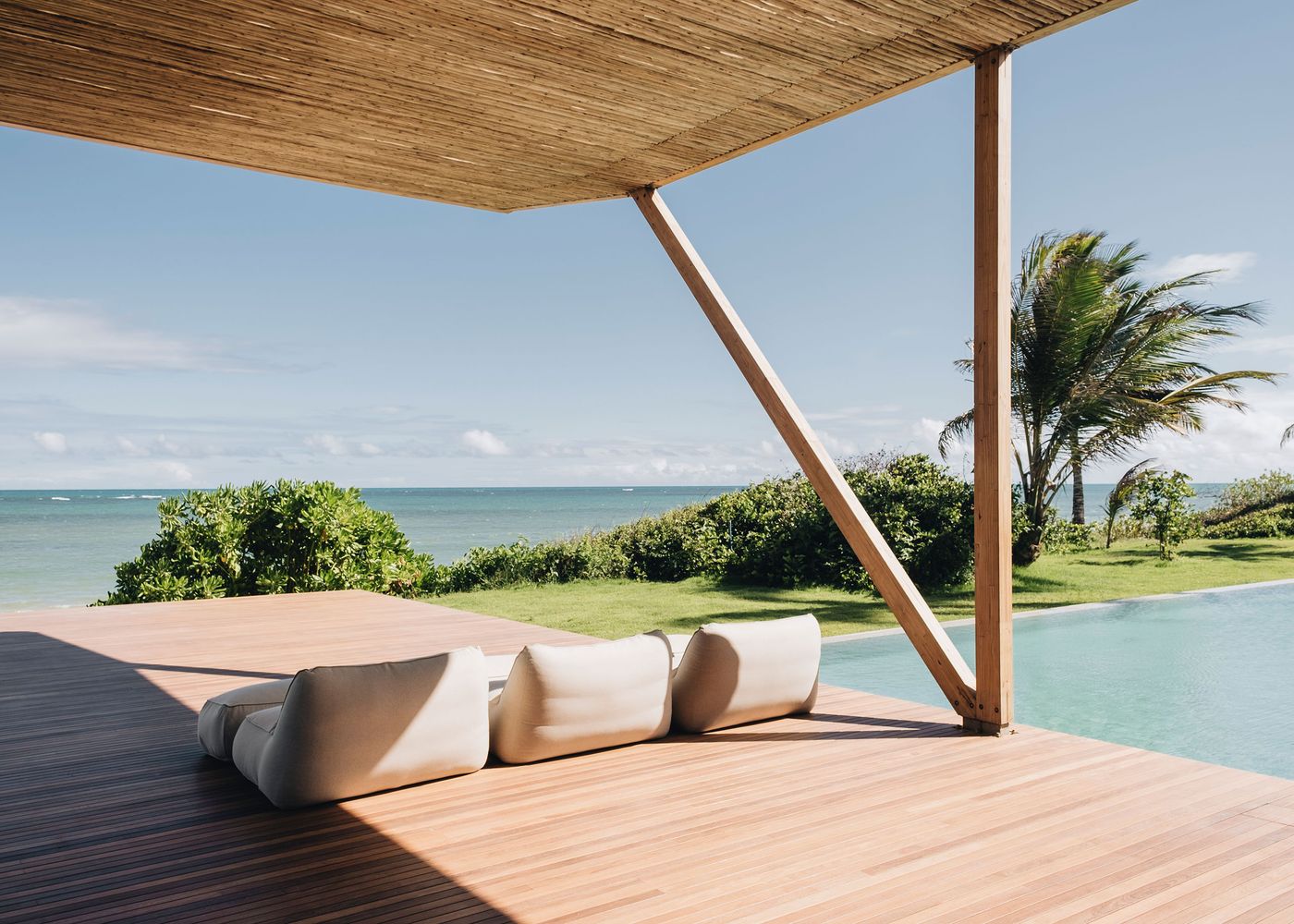 A serene outdoor patio with soft, modern seating overlooking a tranquil sea, framed by lush greenery and a clear blue sky.