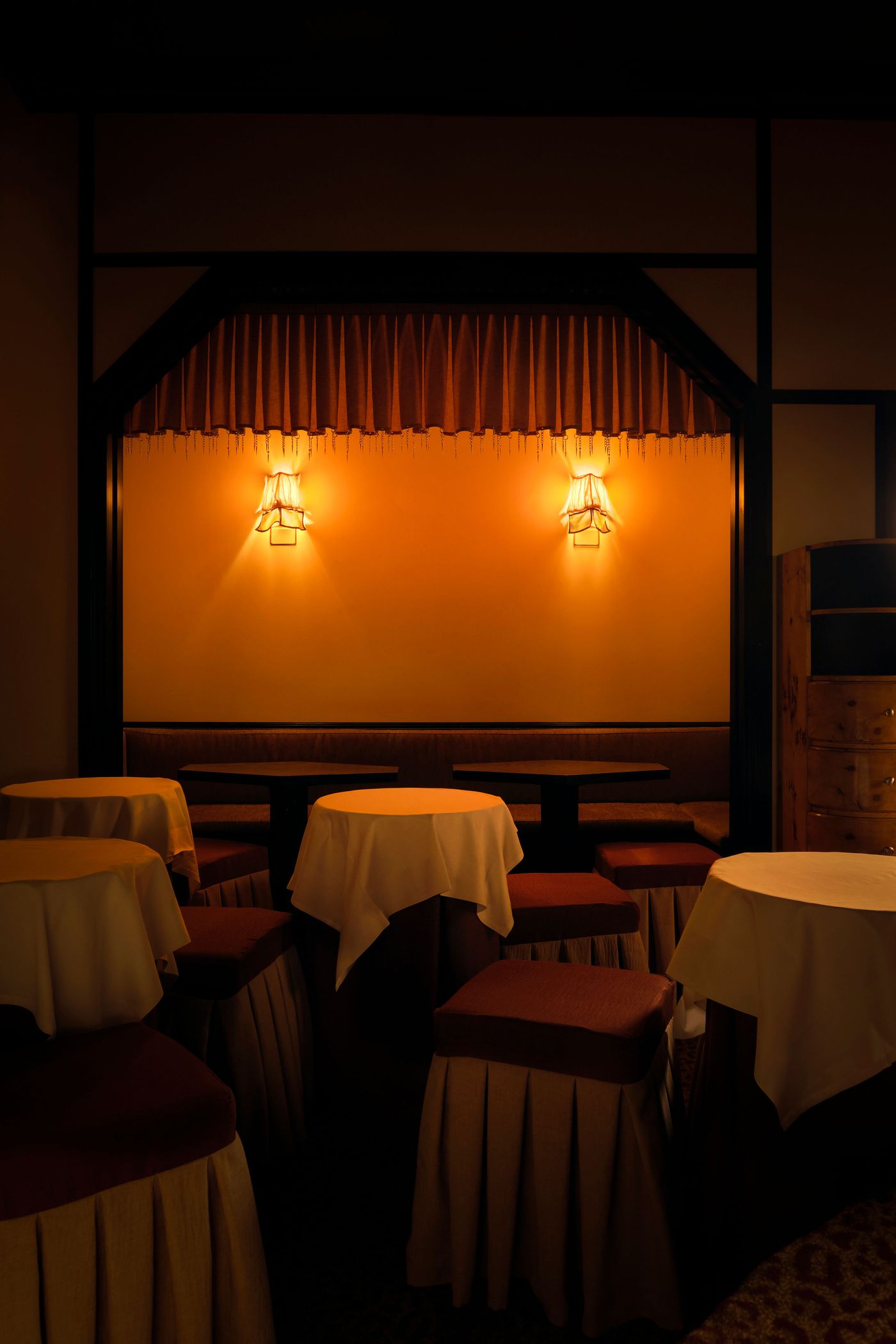 An inviting, dimly lit corner of The Roses of Elagabalus, showcasing an alcove framed by dark wood and soft, pleated drapes. Two ornate sconces with distinctive shades emit a warm, golden light onto the muted yellow wall. Small, white-draped round tables and skirted red stools create intimate seating arrangements, characteristic of a sophisticated retro lounge.