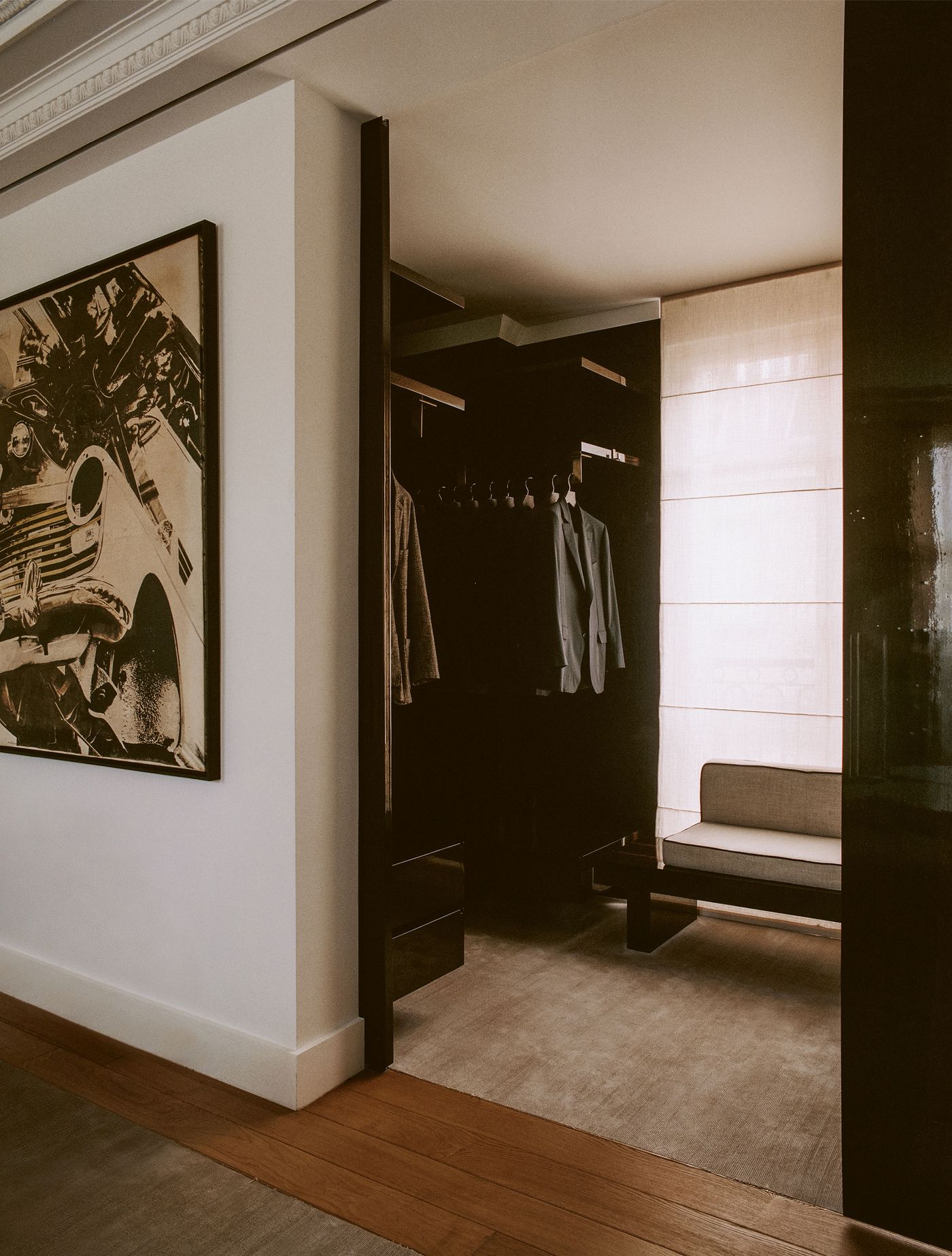 A sophisticated dressing area, partly obscured by a doorway, features dark custom cabinetry for clothing storage, exuding a minimalist yet luxurious feel. A light-toned upholstered bench provides a seating option, contrasted by a large black and white framed artwork of a classic car on an adjacent wall. A sophisticated dressing area, partly obscured by a doorway, features dark custom cabinetry for clothing storage, exuding a minimalist yet luxurious feel. A light-toned upholstered bench provides a seating option, contrasted by a large black and white framed artwork of a classic car on an adjacent wall.