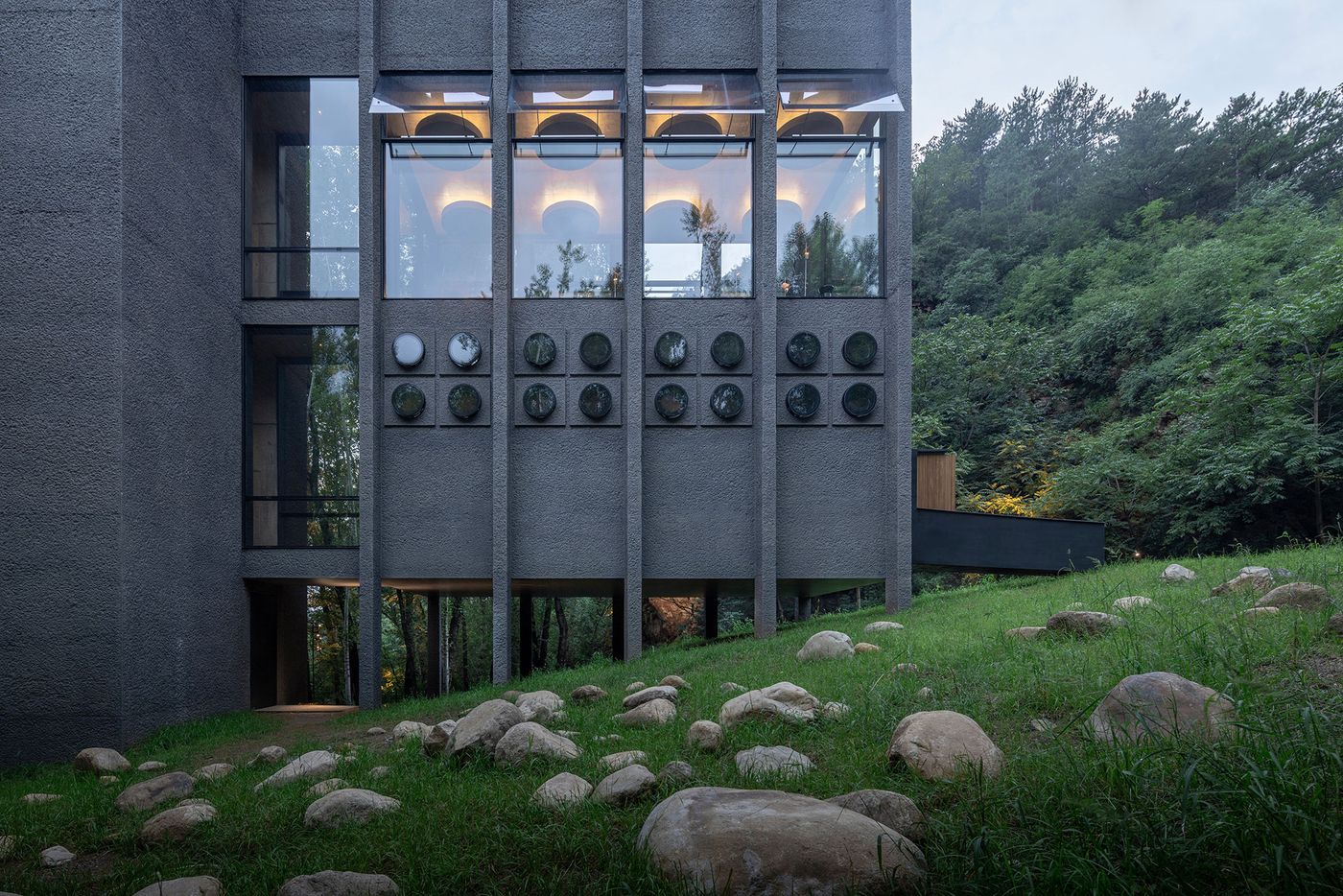 Modern gray building with large windows, circular mirrors, and surrounded by greenery and rocks, blending nature with architecture.
