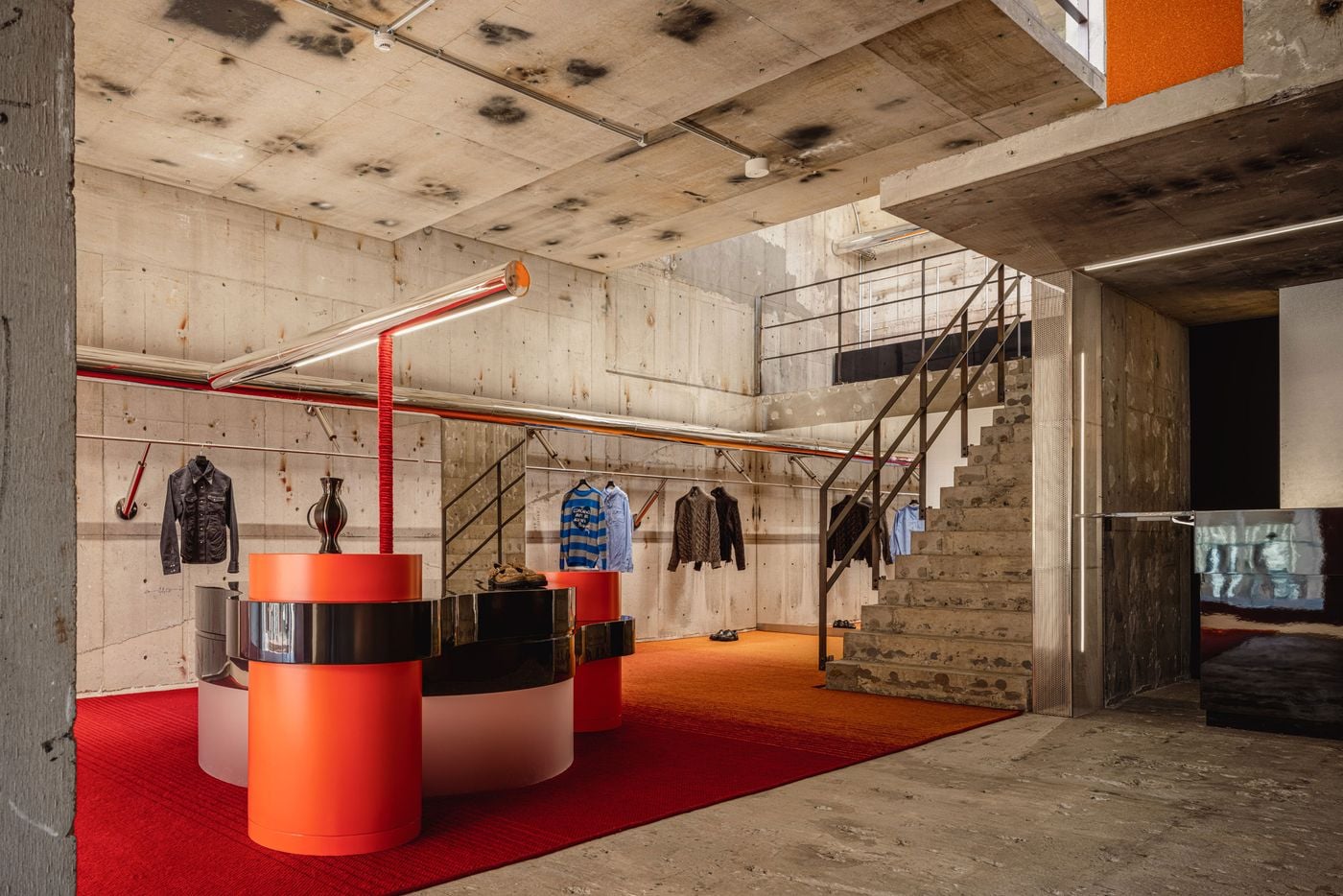 The ground floor juxtaposes exposed, heavily marked concrete with a vivid red-orange floor and sculptural display furniture. Glossy black bands wrap cylindrical orange and frosted bases, while polished chrome rails trace the walls overhead. A raw concrete staircase rises beside the composition, reinforcing the space’s industrial character.