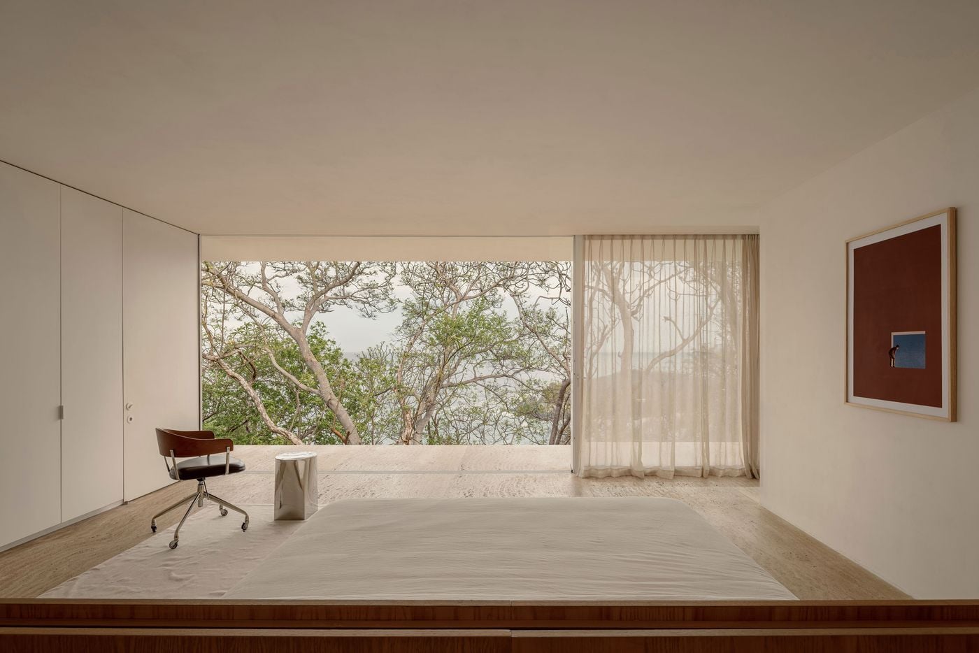 Fully glazed on one side, this bedroom looks straight into the tree canopy, with glimpses of the sea between the branches. Sheer linen curtains, a low bed, a polished steel stool and a leather swivel chair sit on pale paving, while a framed oxblood artwork with a tiny figure at a blue window adds a note of wit. Fully glazed on one side, this bedroom looks straight into the tree canopy, with glimpses of the sea between the branches. Sheer linen curtains, a low bed, a polished steel stool and a leather swivel chair sit on pale paving, while a framed oxblood artwork with a tiny figure at a blue window adds a note of wit.