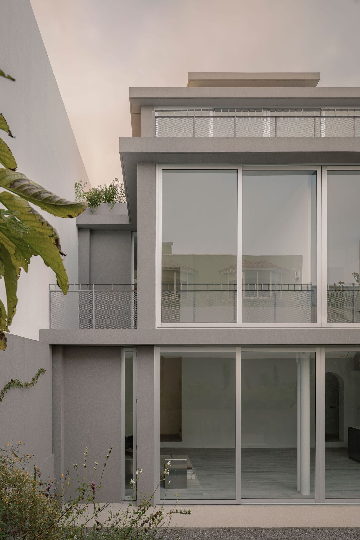 Seen from the garden, Casa Tlaloc’s rear elevation layers expansive glazing, grey structural frames and metal-mesh terraces into a precise vertical composition. Vegetation in the foreground softens the architecture, while reflections of neighbouring roofs across the large windows subtly situate the house within its residential surroundings. Seen from the garden, Casa Tlaloc’s rear elevation layers expansive glazing, grey structural frames and metal-mesh terraces into a precise vertical composition. Vegetation in the foreground softens the architecture, while reflections of neighbouring roofs across the large windows subtly situate the house within its residential surroundings.