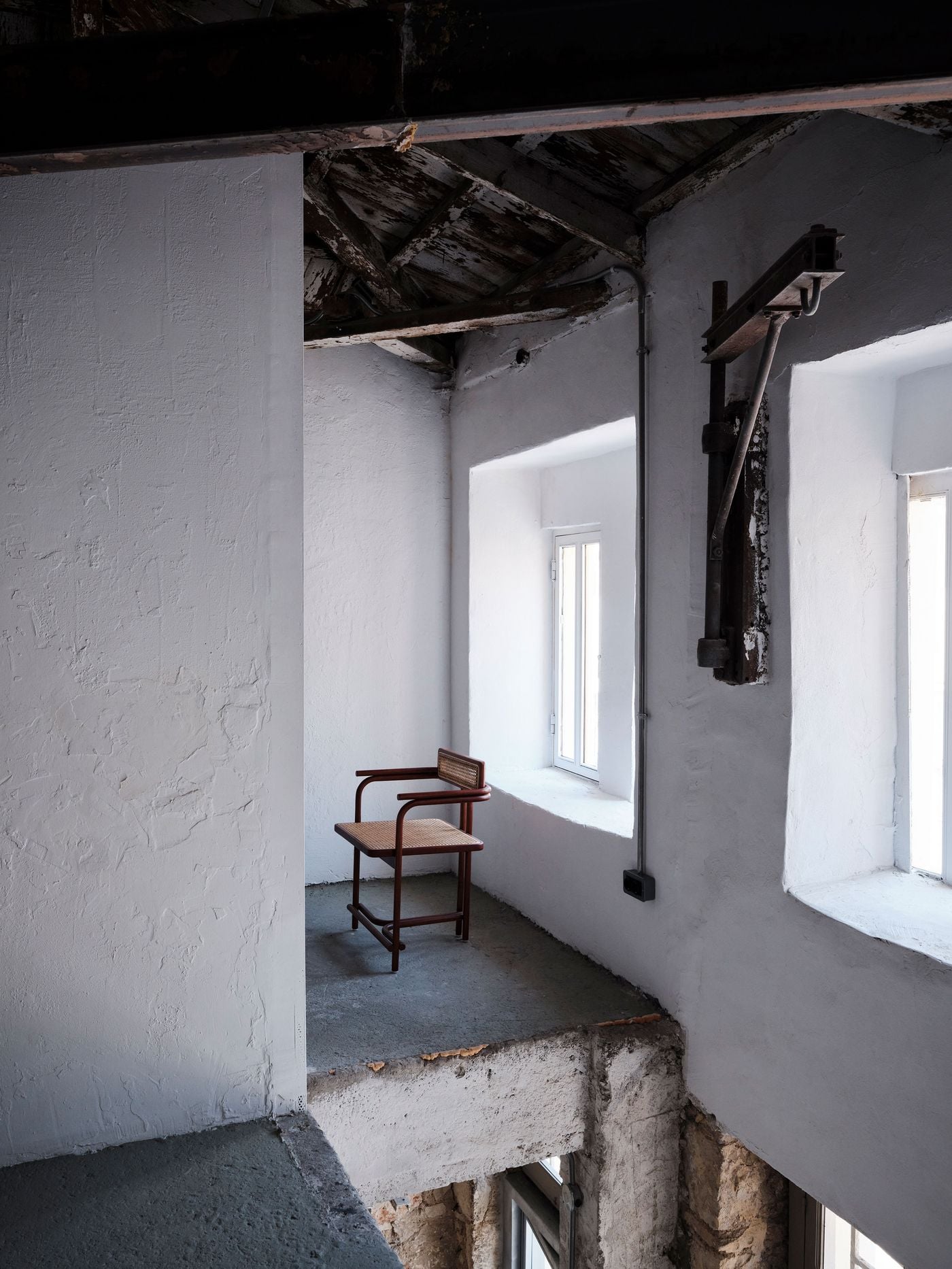 Weathered roof timbers, textured white walls and deep window reveals define this intimate upper-level space. A timber-and-cane chair provides the only furnishing, while an old mechanical fixture remains mounted beside the window, reinforcing MPNYC’s decision to retain fragments from the building’s successive industrial lives. Weathered roof timbers, textured white walls and deep window reveals define this intimate upper-level space. A timber-and-cane chair provides the only furnishing, while an old mechanical fixture remains mounted beside the window, reinforcing MPNYC’s decision to retain fragments from the building’s successive industrial lives.
