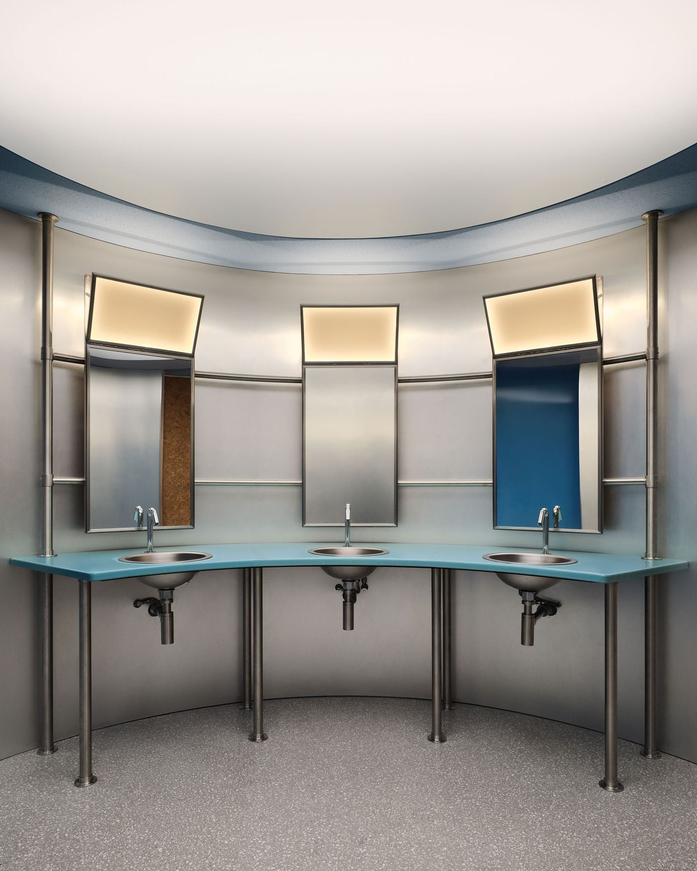 A curved wash area in the Ken Club’s locker room pairs a powder-blue countertop with brushed metal walls, exposed stainless-steel basins and pipework. Three illuminated mirrors tilt slightly forward beneath a large circular ceiling light, giving the space a playful, almost spaceship-like character. A curved wash area in the Ken Club’s locker room pairs a powder-blue countertop with brushed metal walls, exposed stainless-steel basins and pipework. Three illuminated mirrors tilt slightly forward beneath a large circular ceiling light, giving the space a playful, almost spaceship-like character.