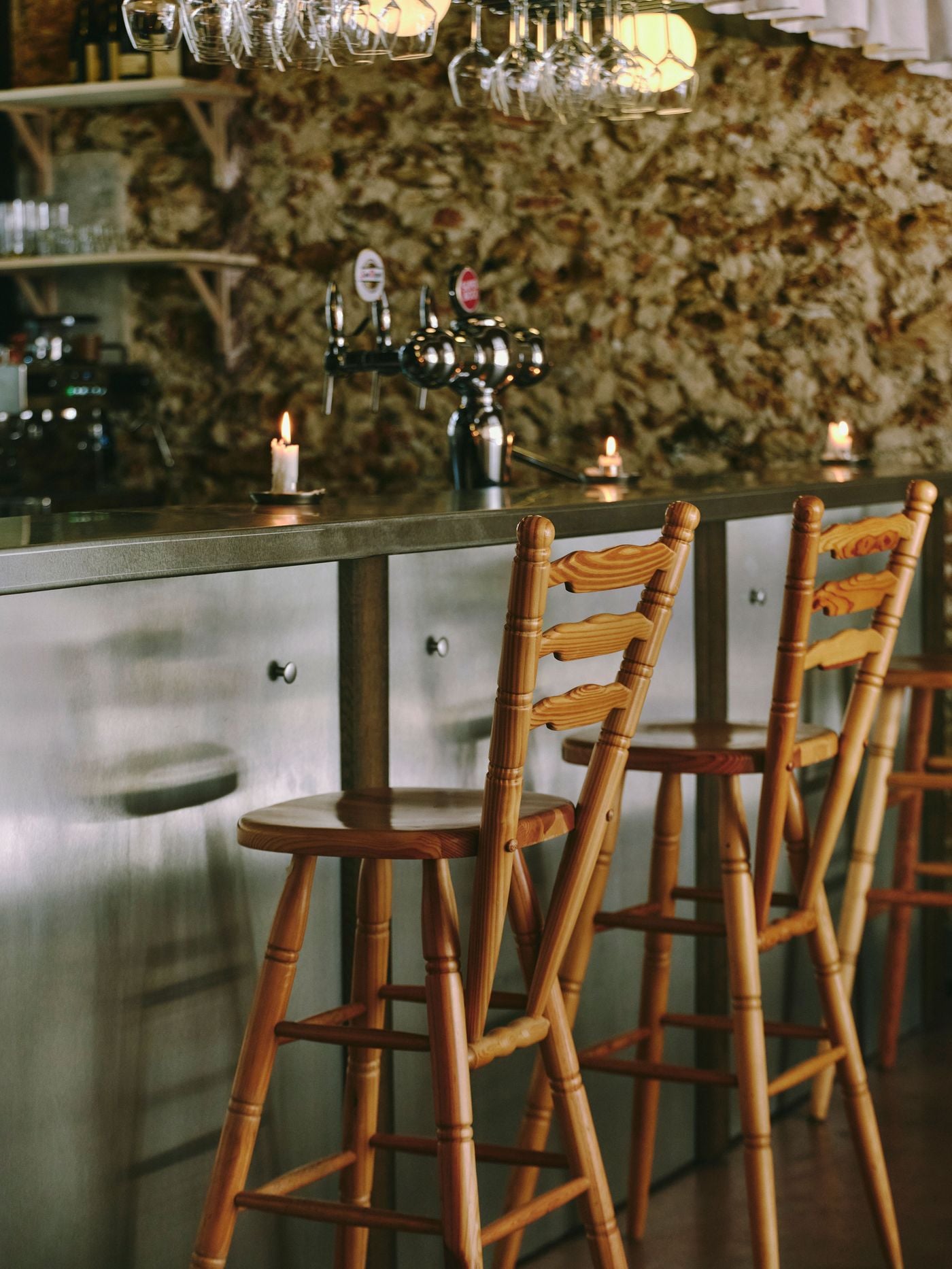 Pine ladder-back stools with carved rails and turned legs pull up to the brushed-steel bar at SaltoBar in Paris. Behind them, chrome beer taps and stubby candles stand against a rough rubblestone wall. Rows of inverted wine glasses hang from racks overhead, catching the glow of spherical lamps. Pine ladder-back stools with carved rails and turned legs pull up to the brushed-steel bar at SaltoBar in Paris. Behind them, chrome beer taps and stubby candles stand against a rough rubblestone wall. Rows of inverted wine glasses hang from racks overhead, catching the glow of spherical lamps.