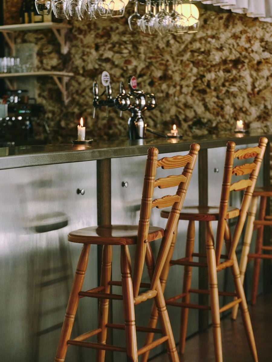 Pine ladder-back stools with carved rails and turned legs pull up to the brushed-steel bar at SaltoBar in Paris. Behind them, chrome beer taps and stubby candles stand against a rough rubblestone wall. Rows of inverted wine glasses hang from racks overhead, catching the glow of spherical lamps.