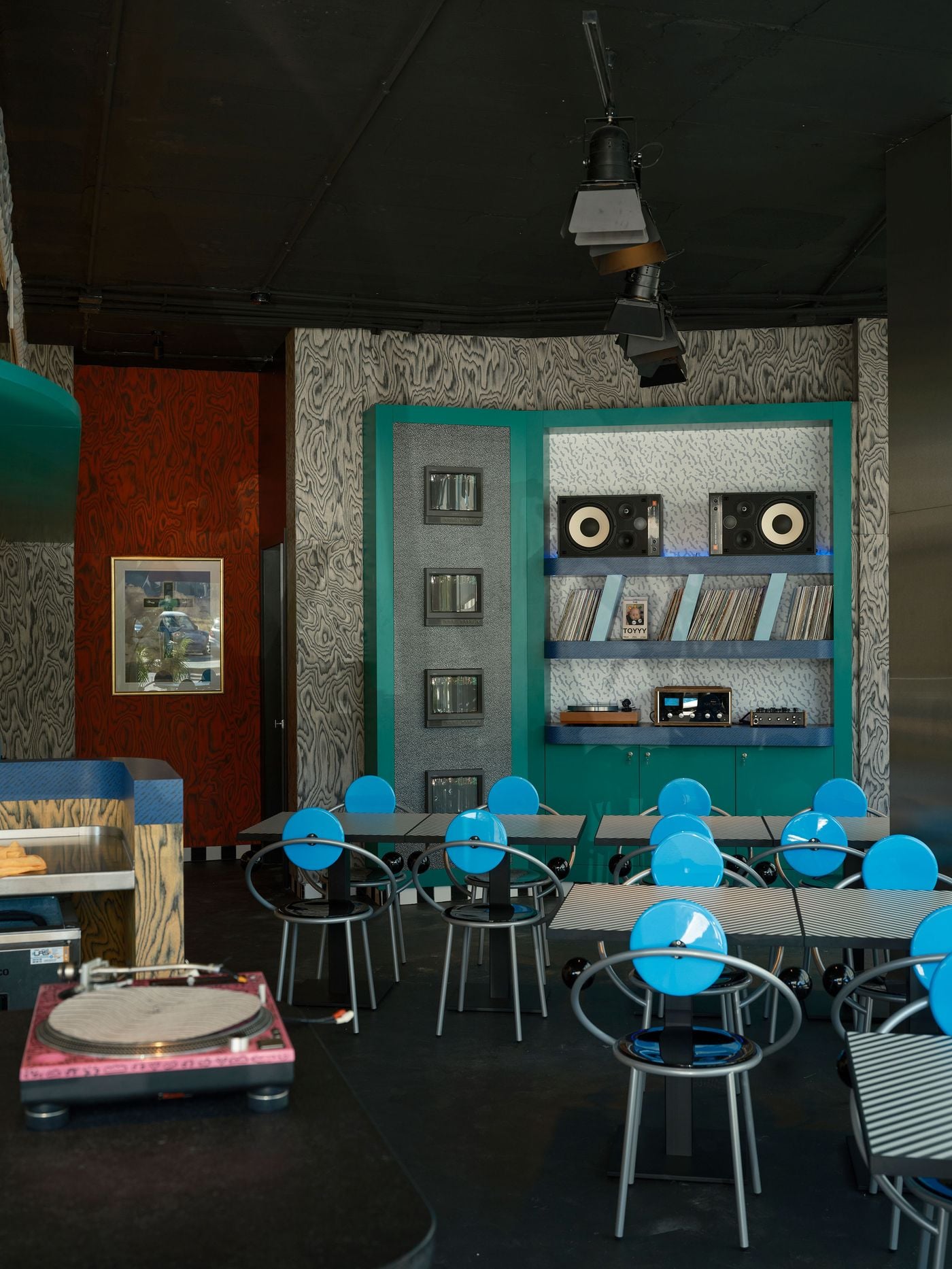 A pink turntable in the foreground sets the tone for the dining room at Seth & Manni's, where Michele De Lucchi’s blue disc-backed First chairs gather around striped tables. Beyond, the teal media wall of monitors, speakers and records meets grey and rust-red wood-grain walls. Theatrical spotlights hang from the black ceiling. A pink turntable in the foreground sets the tone for the dining room at Seth & Manni's, where Michele De Lucchi’s blue disc-backed First chairs gather around striped tables. Beyond, the teal media wall of monitors, speakers and records meets grey and rust-red wood-grain walls. Theatrical spotlights hang from the black ceiling.