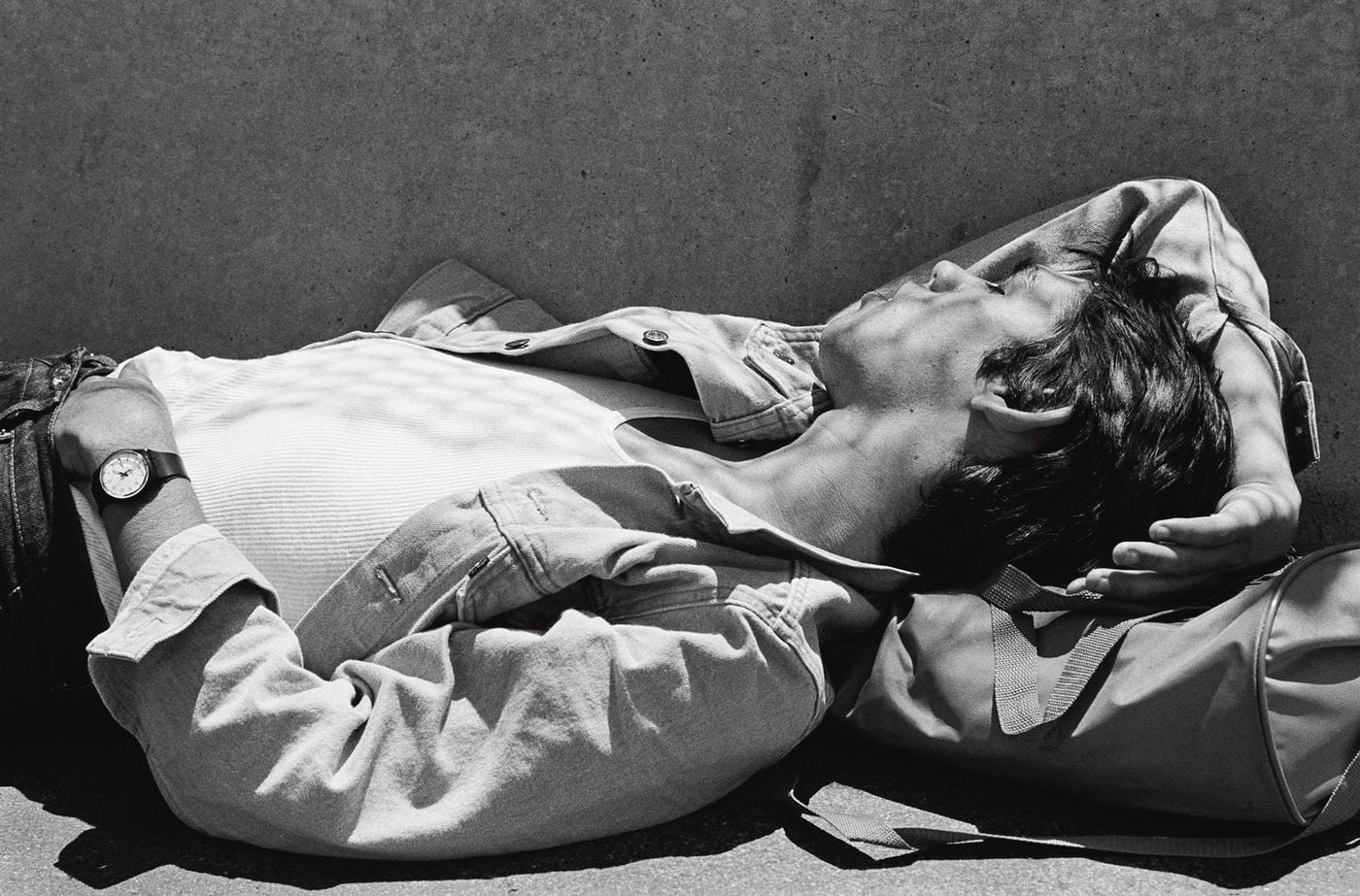 A young man dozes in the sun on a concrete ledge, his head resting on a sports holdall, one arm curled behind his tousled hair. A worn denim jacket falls open over a ribbed white vest, and a wristwatch catches the light, a languid study of downtime from Stanley Stellar's Don't Worry Boys. A young man dozes in the sun on a concrete ledge, his head resting on a sports holdall, one arm curled behind his tousled hair. A worn denim jacket falls open over a ribbed white vest, and a wristwatch catches the light, a languid study of downtime from Stanley Stellar's Don't Worry Boys.