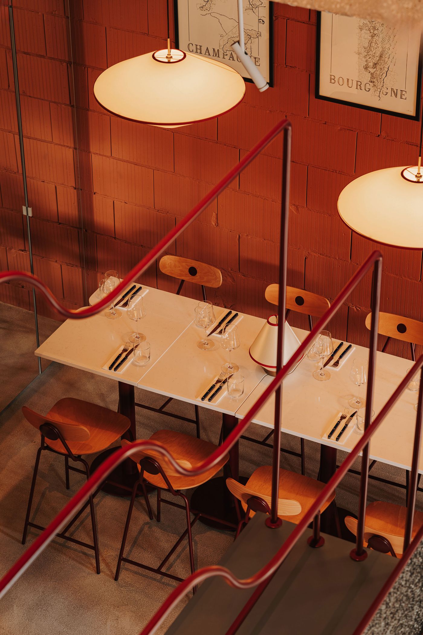 Viewed from the staircase, a dining area at WIN wine bar combines neatly set white tables, warm timber chairs and terracotta-coloured masonry walls. Sculptural pendant lights cast a soft glow over the setting, while sinuous burgundy metal balustrades sweep across the foreground, their fluid profiles contrasting with the space's rectilinear architecture. Viewed from the staircase, a dining area at WIN wine bar combines neatly set white tables, warm timber chairs and terracotta-coloured masonry walls. Sculptural pendant lights cast a soft glow over the setting, while sinuous burgundy metal balustrades sweep across the foreground, their fluid profiles contrasting with the space's rectilinear architecture.