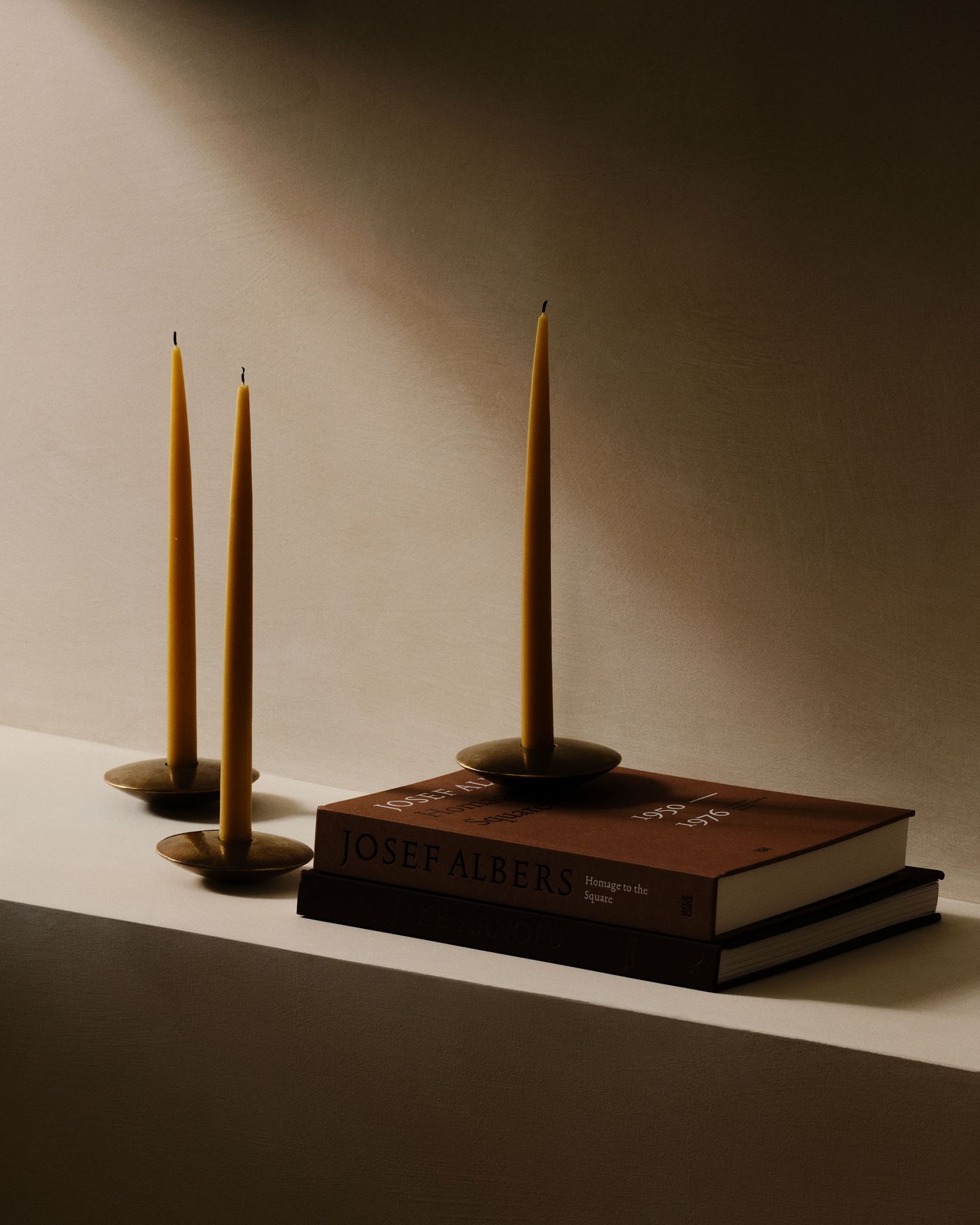 A minimalist scene featuring three yellow candles on brass holders atop stacked art books, with soft lighting and textured wall backdrop.