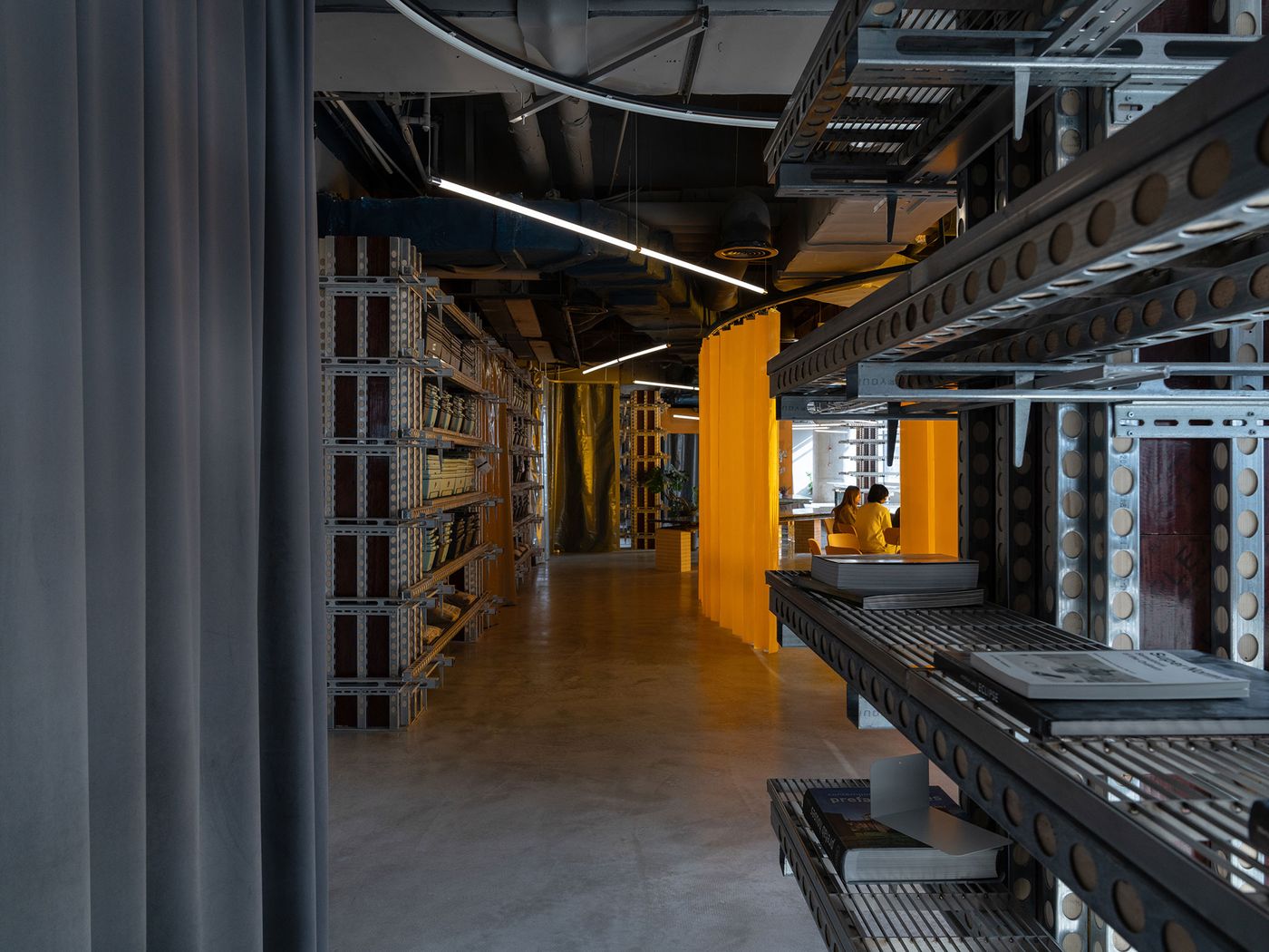 This interior perspective of 889 GLO Art Space showcases its elongated layout, featuring an extensive industrial shelving unit displaying books and objects. A vivid yellow curtain introduces a burst of color, drawing the eye towards a sunlit area where people gather, seamlessly blending the raw brutalist structure with vibrant, inviting communal zones. This interior perspective of 889 GLO Art Space showcases its elongated layout, featuring an extensive industrial shelving unit displaying books and objects. A vivid yellow curtain introduces a burst of color, drawing the eye towards a sunlit area where people gather, seamlessly blending the raw brutalist structure with vibrant, inviting communal zones.
