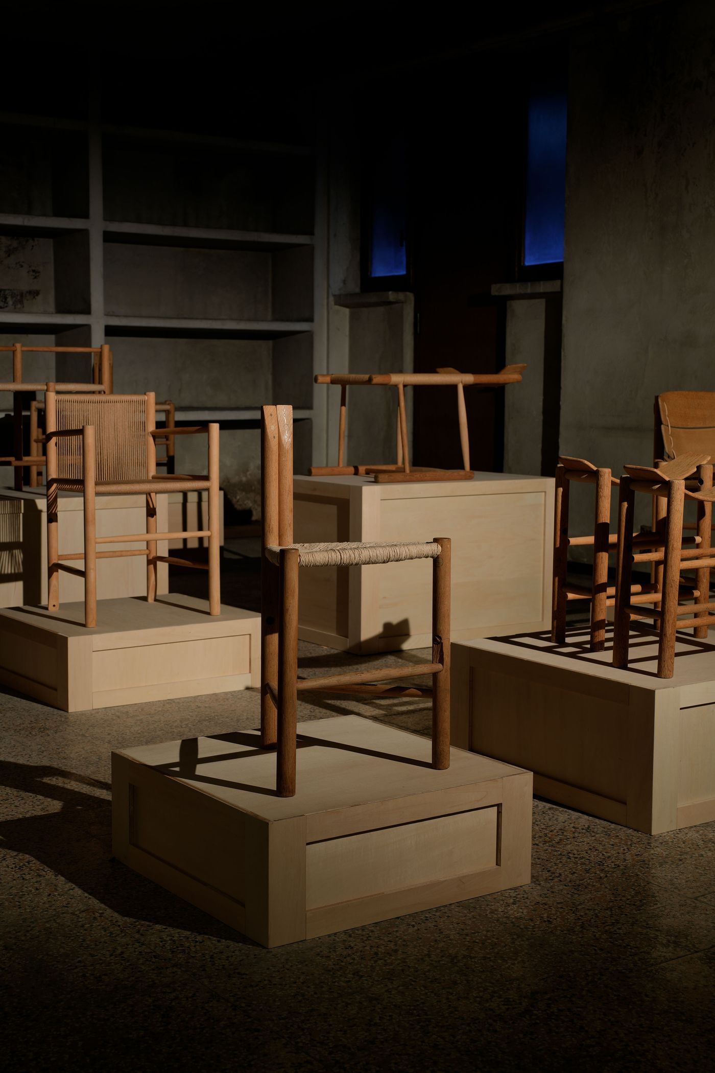 A collection of wooden chairs on pedestals, illuminated in a dark space, highlighting their unique designs and materials.