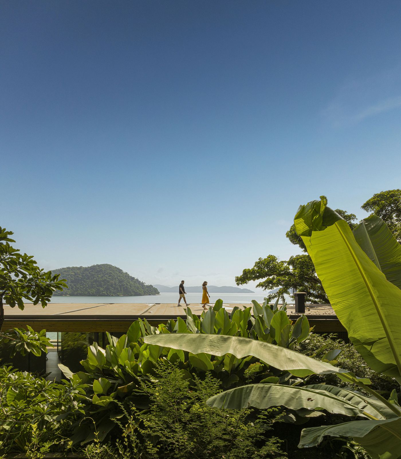 Two individuals walk along the elevated walkway of the modernist residence, seemingly floating above a dense sea of vibrant green tropical plants. Beyond, the tranquil ocean stretches to distant, hazy mountains, highlighting the architectural strategy of elevating the living spaces to embrace panoramic views and a biophilic aesthetic.