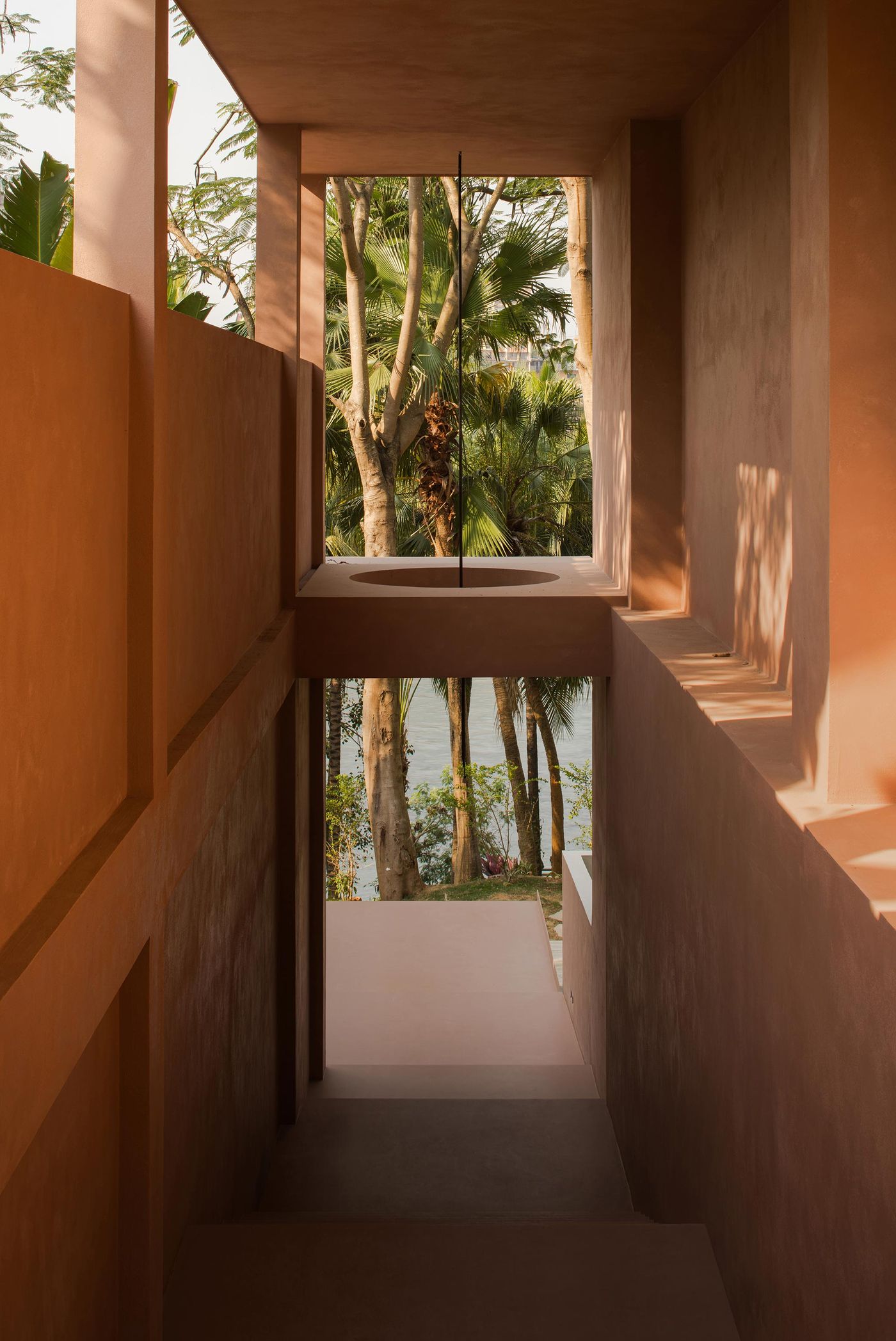 A sunlit corridor with terracotta walls, leading to a view of lush trees and water through a large, framed window. A sunlit corridor with terracotta walls, leading to a view of lush trees and water through a large, framed window.
